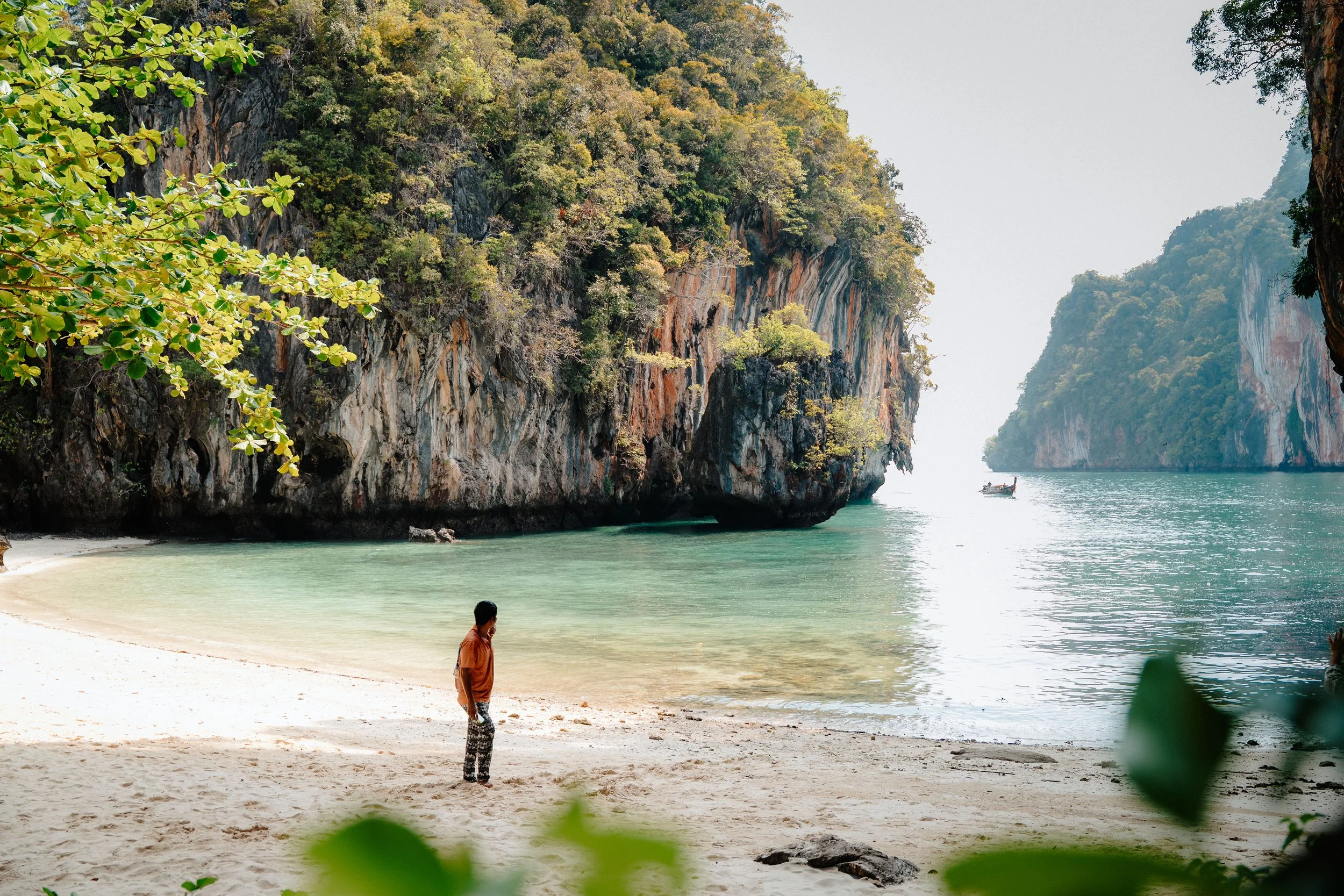 A beach with white sand, surrounded by lush green hills and tall cliffs, with calm turquoise water. Two people are visible, one standing near the shoreline and another walking along the beach, with a small boat in the water.