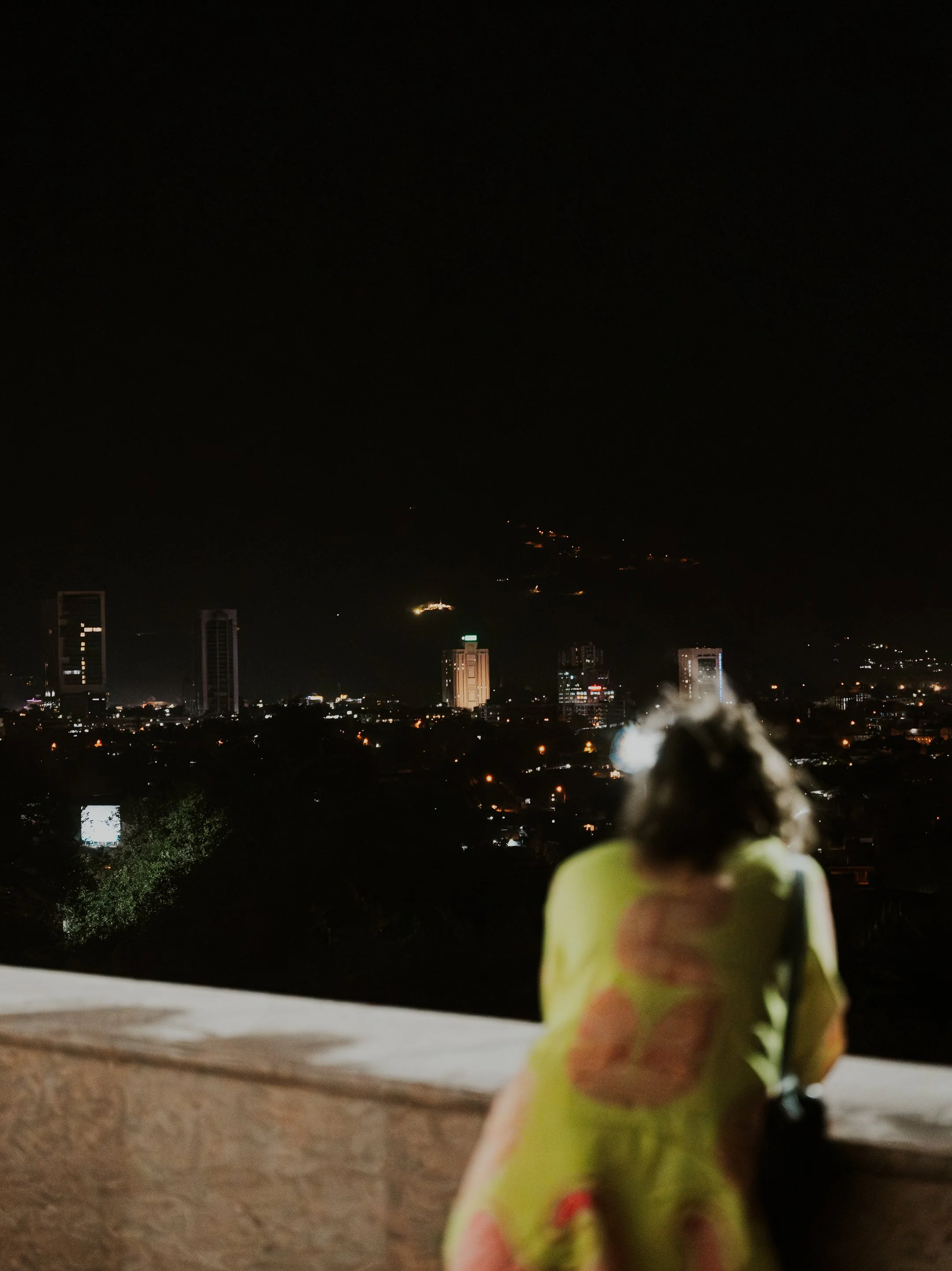 A small plush toy car with a colorful floral blanket draped over it, placed on a ledge overlooking a cityscape at night with tall buildings and distant hills.