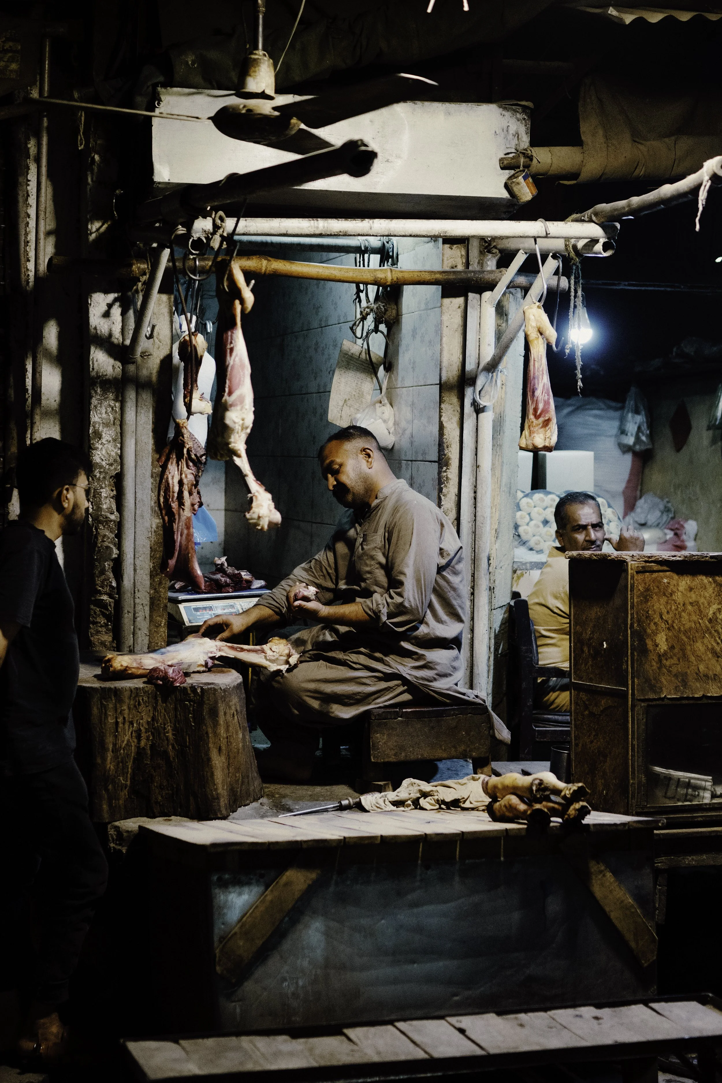 A meat stall in a market with dried meat and hanging cuts, a man preparing meat, and others behind the counter.