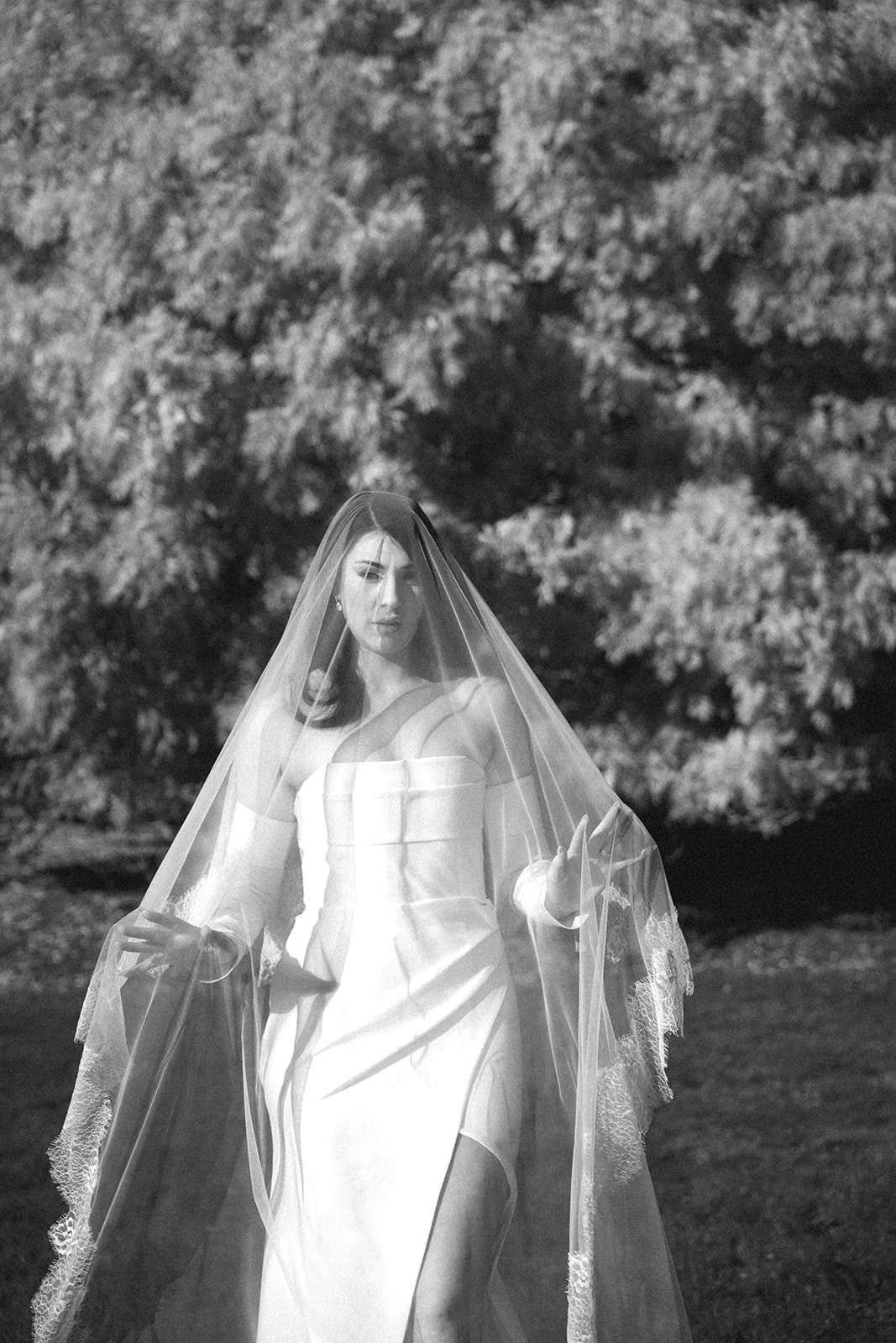 Black and white photo of a woman in a wedding dress and veil standing outdoors with trees in the background.