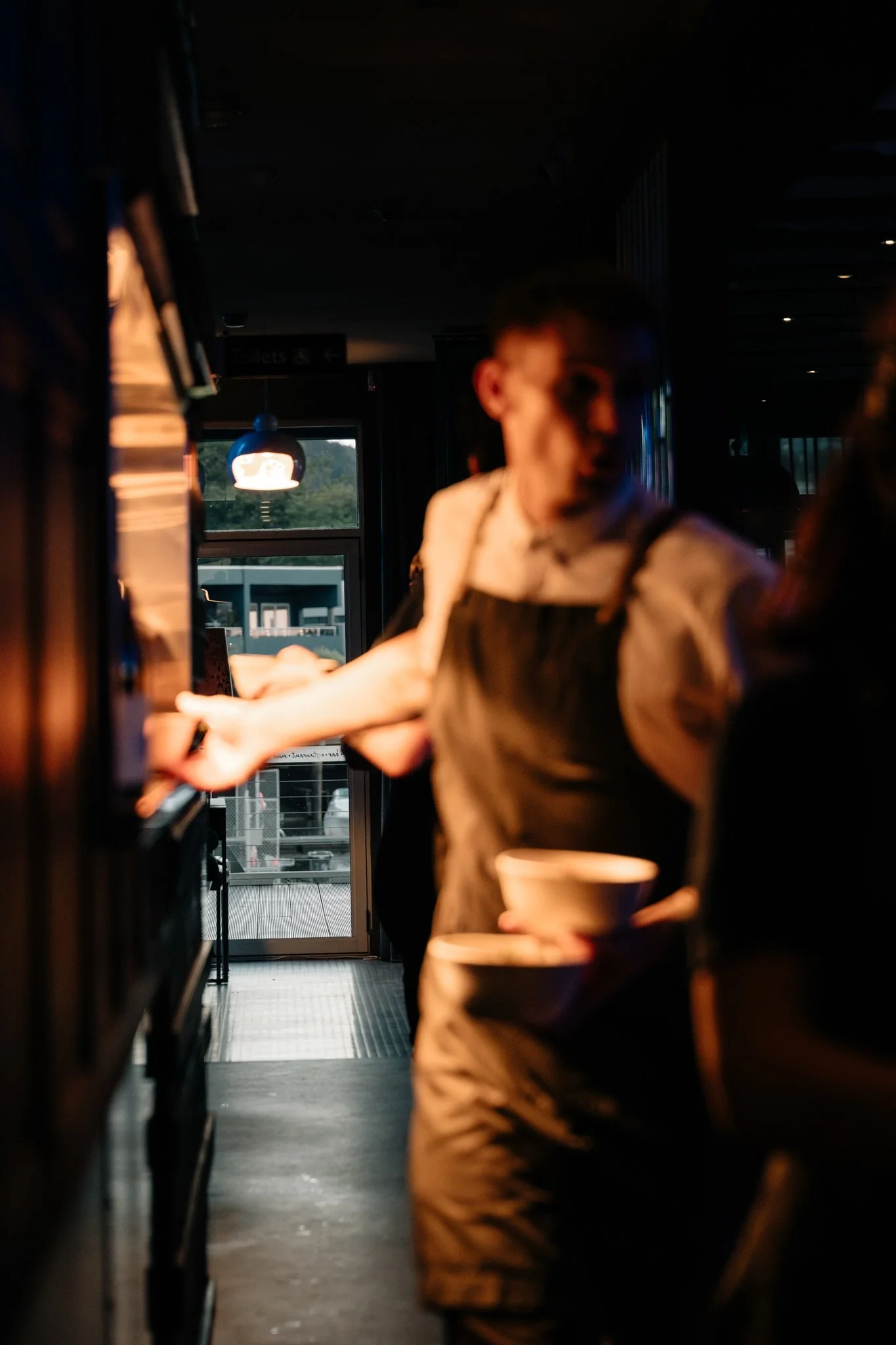 People in a dimly lit coffee shop or café, with one person in focus holding a paper cup, while others are blurred and in motion, with a door and outdoor view of a building and greenery visible in the background.