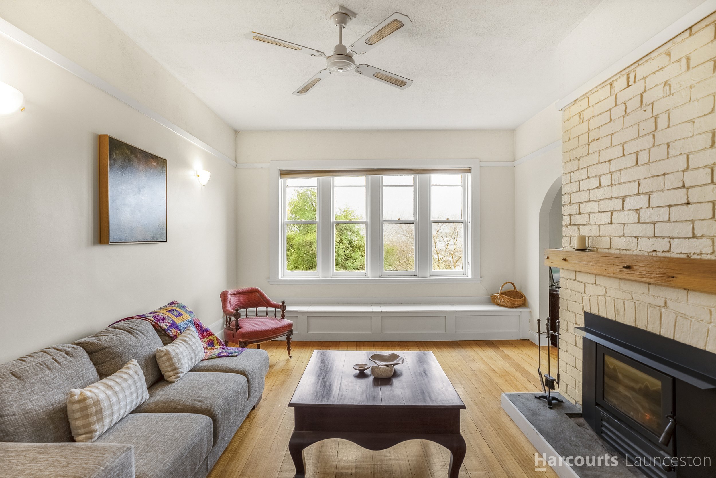 Living room with a beige sofa, a wooden coffee table, a red chair, a fireplace with a brick wall, and a large window with trees outside.