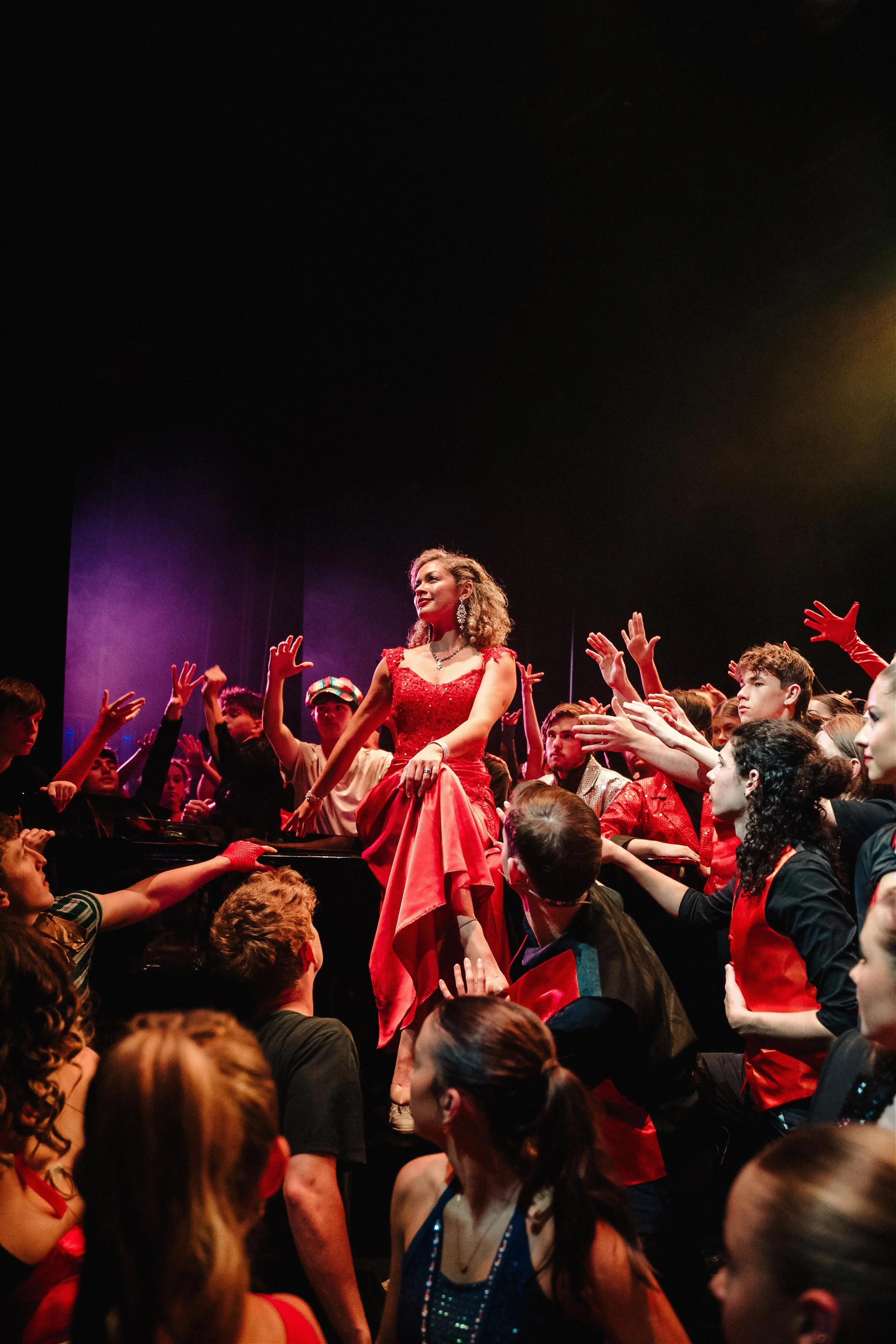 A woman in a red dress standing on a stage surrounded by a crowd of people reaching out to her, with colorful stage lighting.