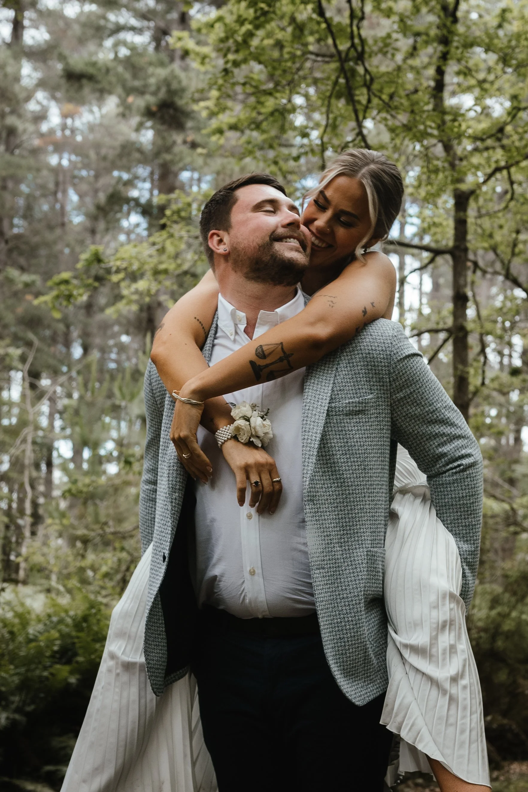 A couple in formal attire sharing a joyful moment outdoors in a forest, with the woman leaning on the man's back and both smiling.