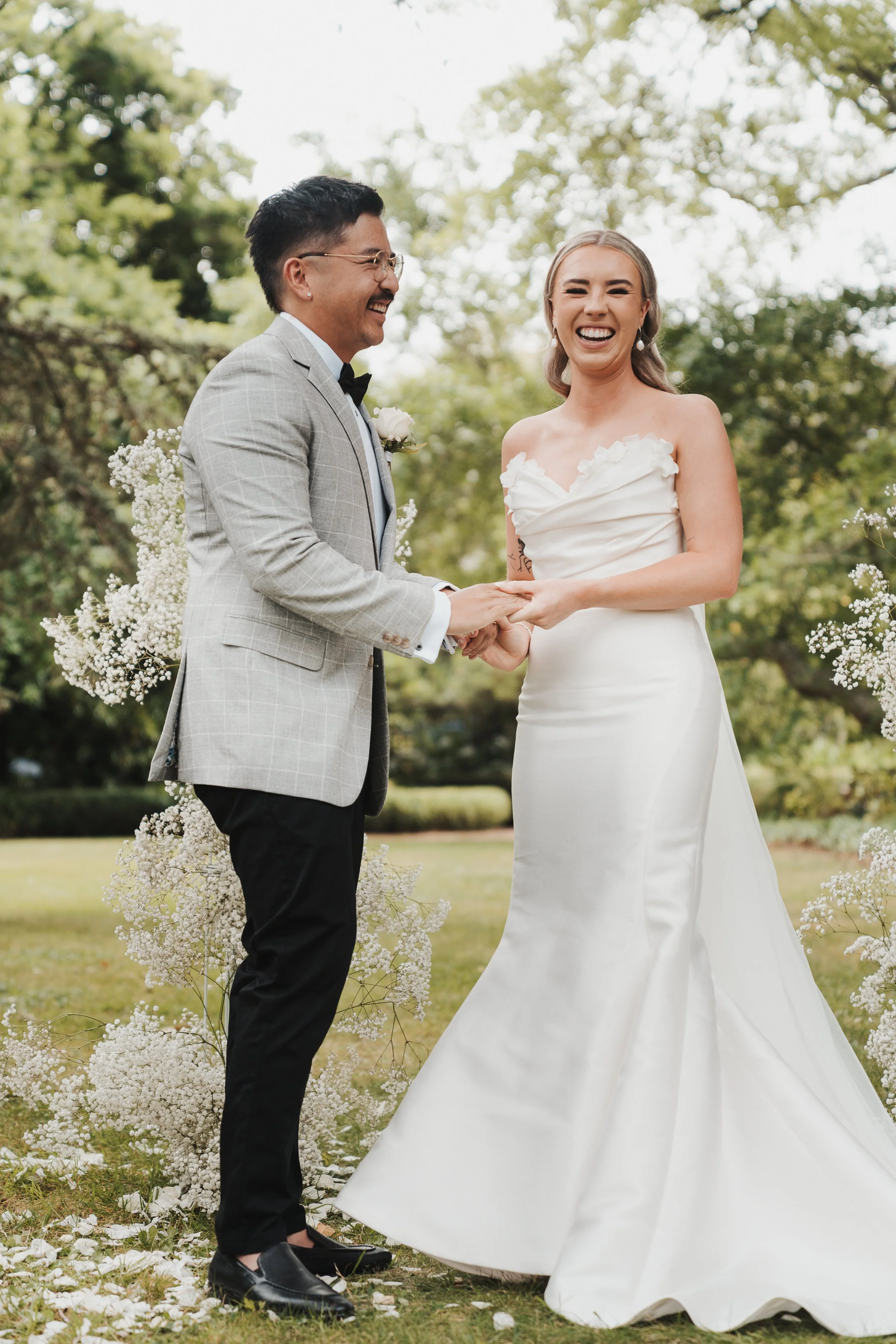 A bride and groom holding hands and smiling during their outdoor wedding ceremony. The bride is wearing a white strapless wedding gown, and the groom is dressed in a light gray suit with a black bow tie. Green trees and white floral arrangements are 