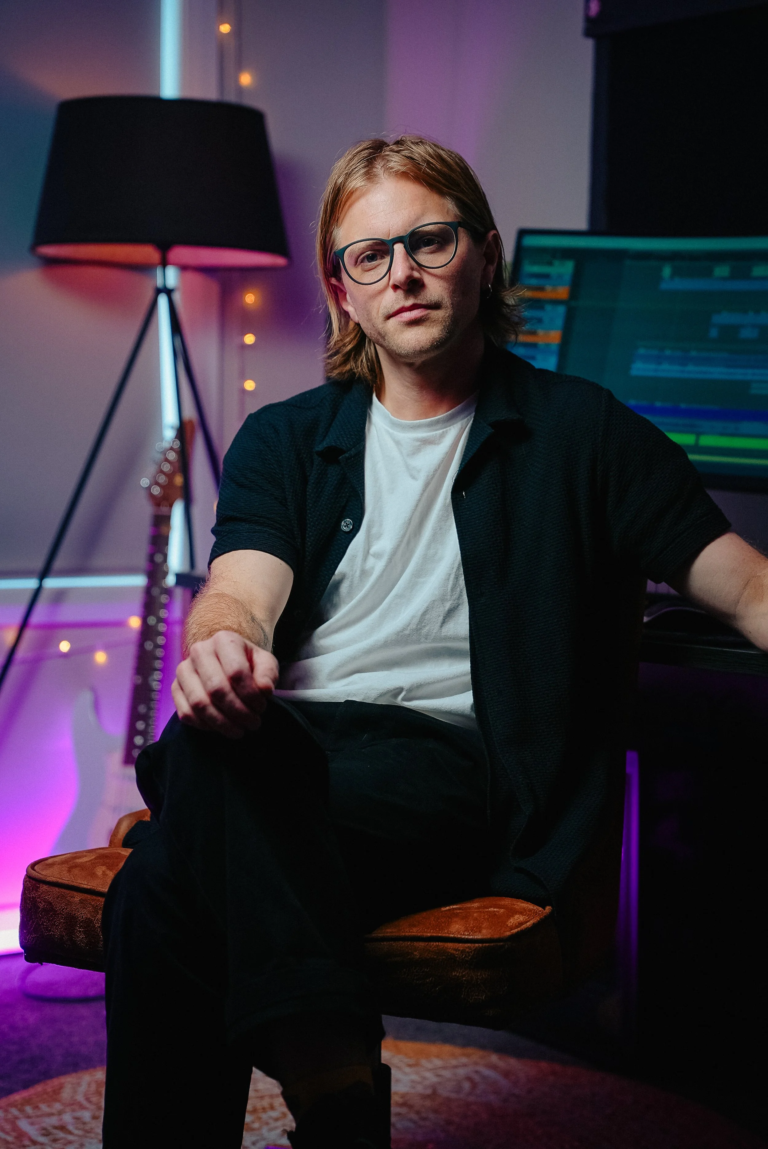 A man with glasses and long hair sitting on a brown chair in a dimly lit room with colorful lights, a computer screen displaying editing software in the background, a tall black lamp, and a guitar visible in the background.