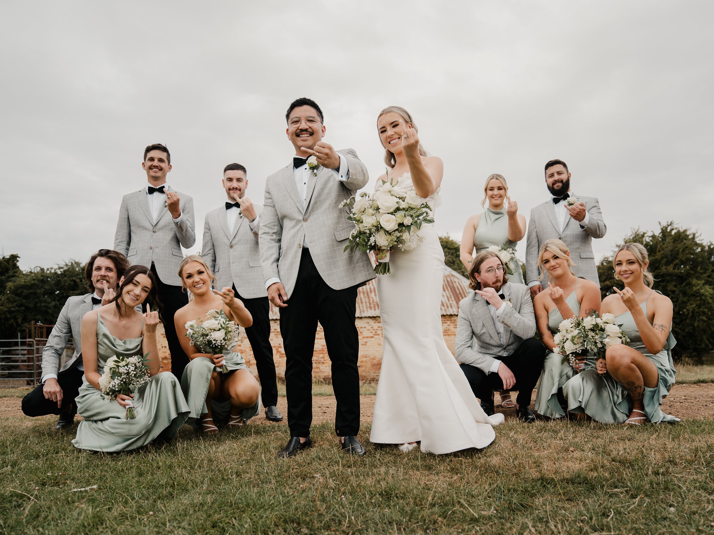 A wedding party outside on grass under a cloudy sky, with people in formal attire making finger heart gestures.