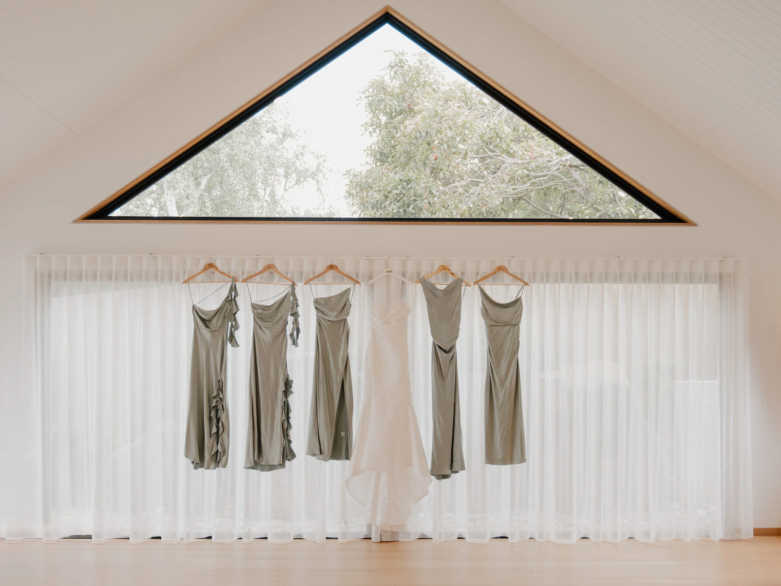 A triangular skylight in a ceiling revealing an outside view of green trees and sky. Below, beige and gray clothing hangs on hangers in front of sheer white curtains.