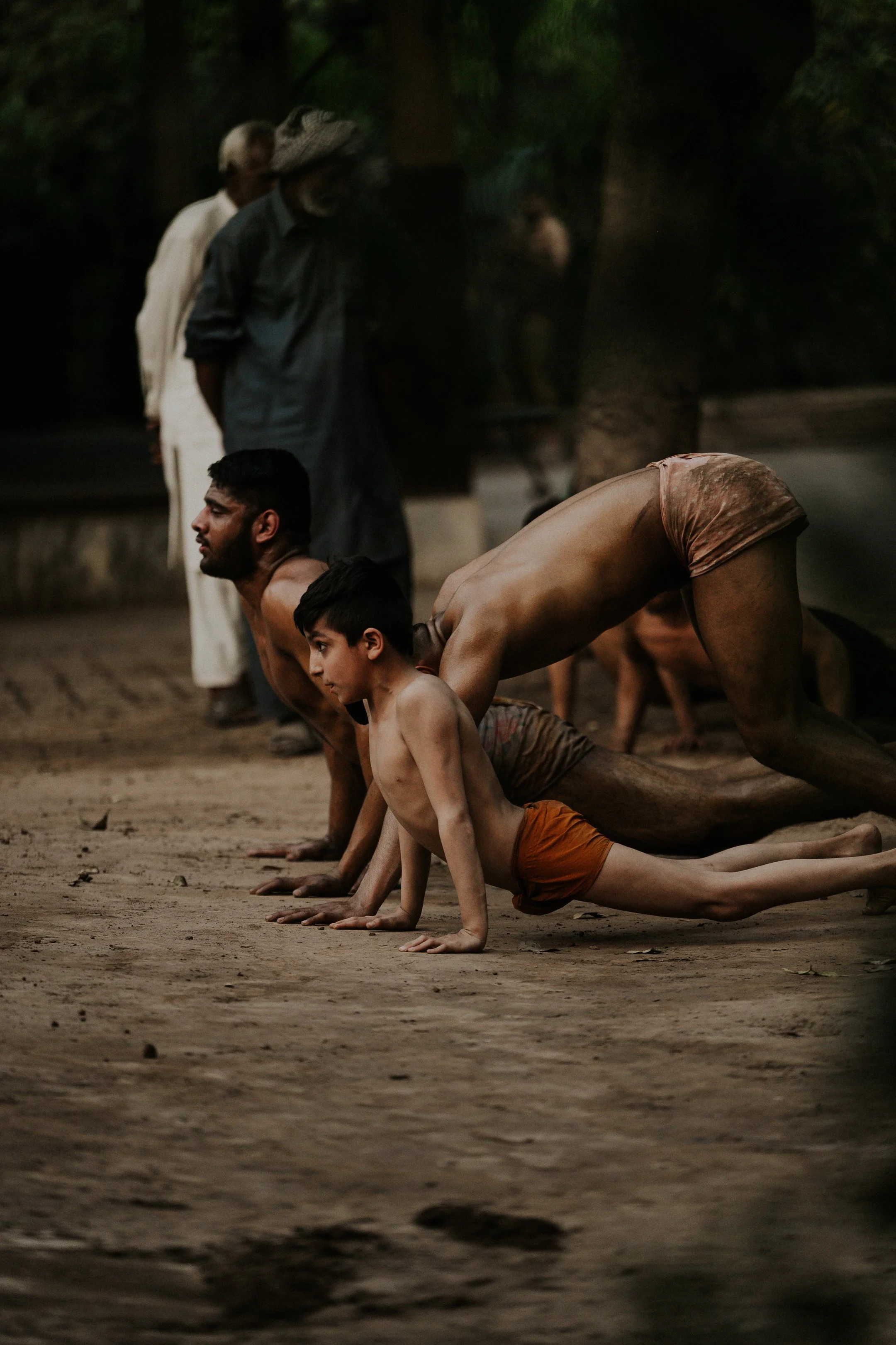 Three children and one adult practicing yoga or stretching outdoors on a dirt ground with trees in the background, during dusk or dawn.
