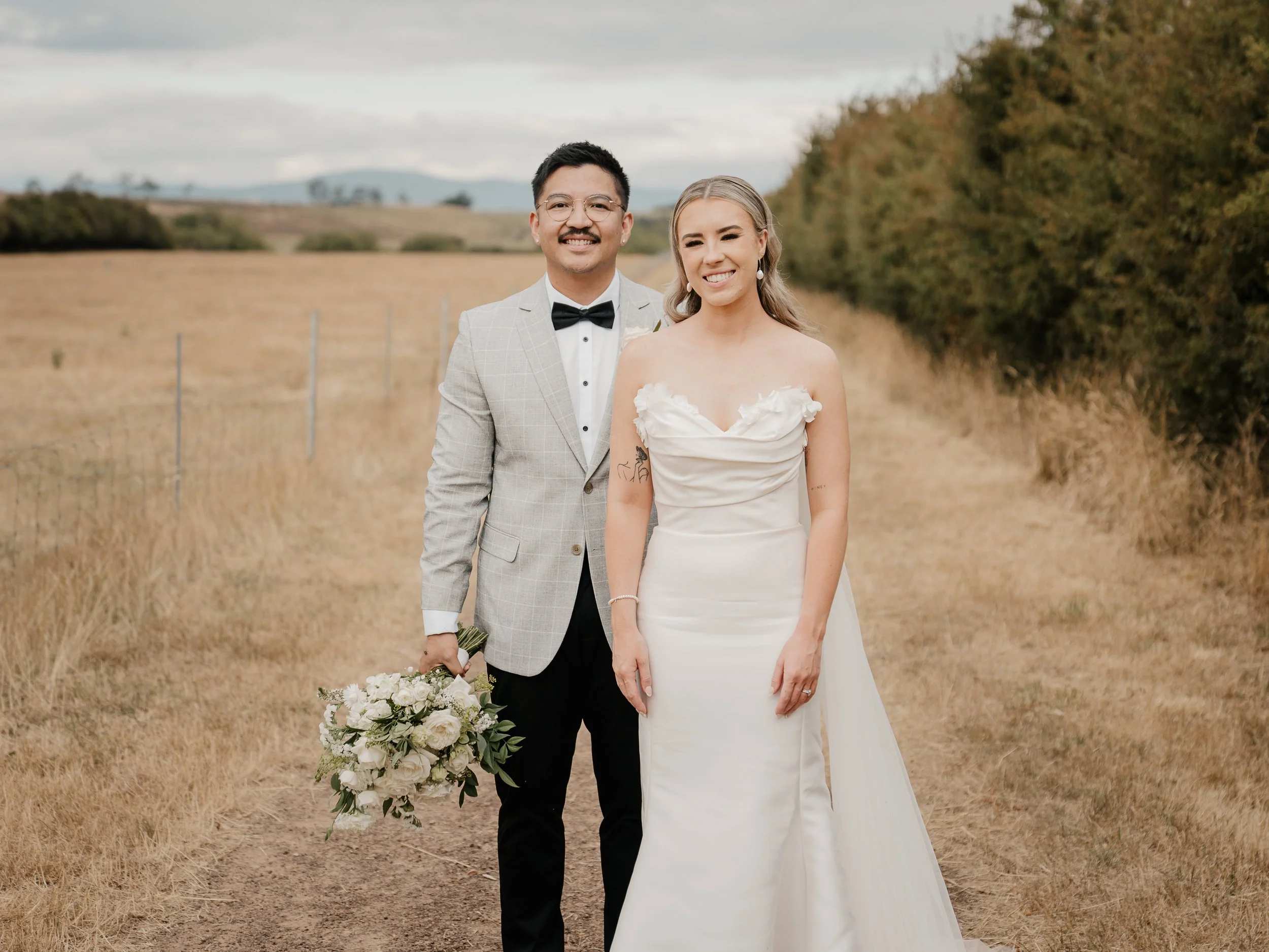 A happy bride and groom standing outdoors in a field with trees and mountains in the distance. The groom is dressed in a light-colored suit with a black bow tie. The bride is wearing a white wedding gown with floral details on the bodice and holding 
