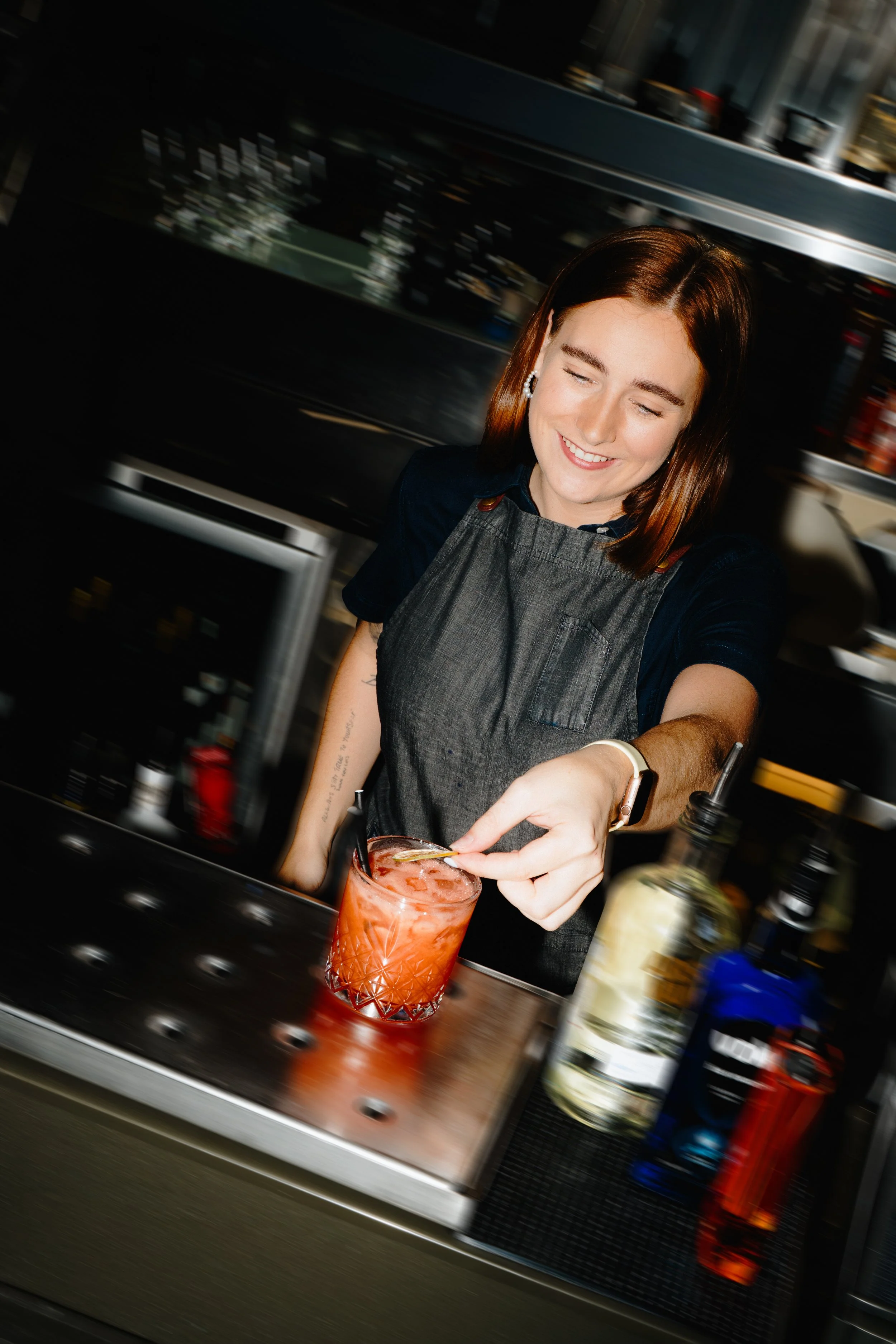 A bartender making a cocktail behind a bar counter, wearing a dark shirt and apron, with bottles of alcohol and bar equipment in the background.