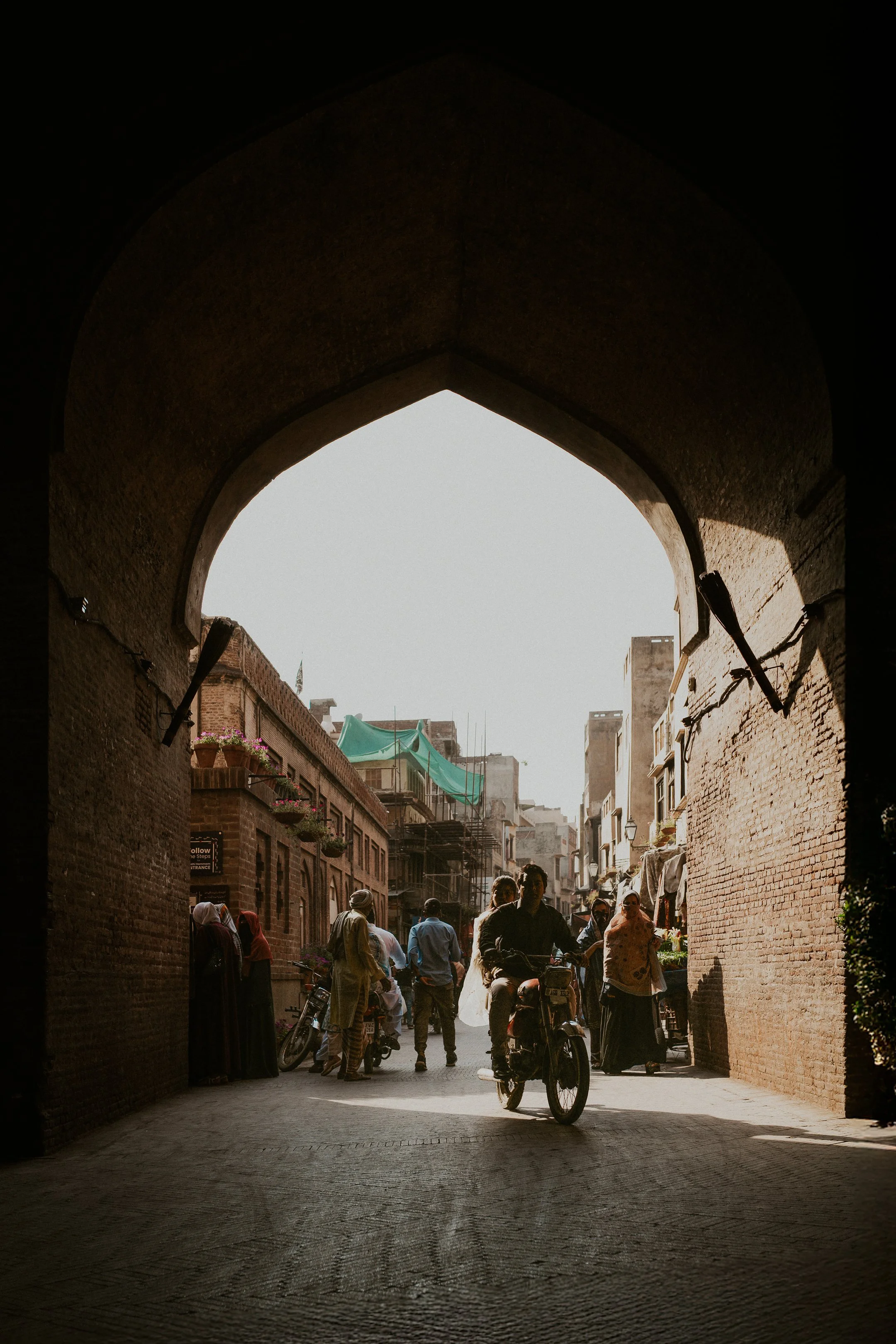 People walking and riding bicycles through an archway in a busy street market area with buildings on either side and a green canopy in the background.