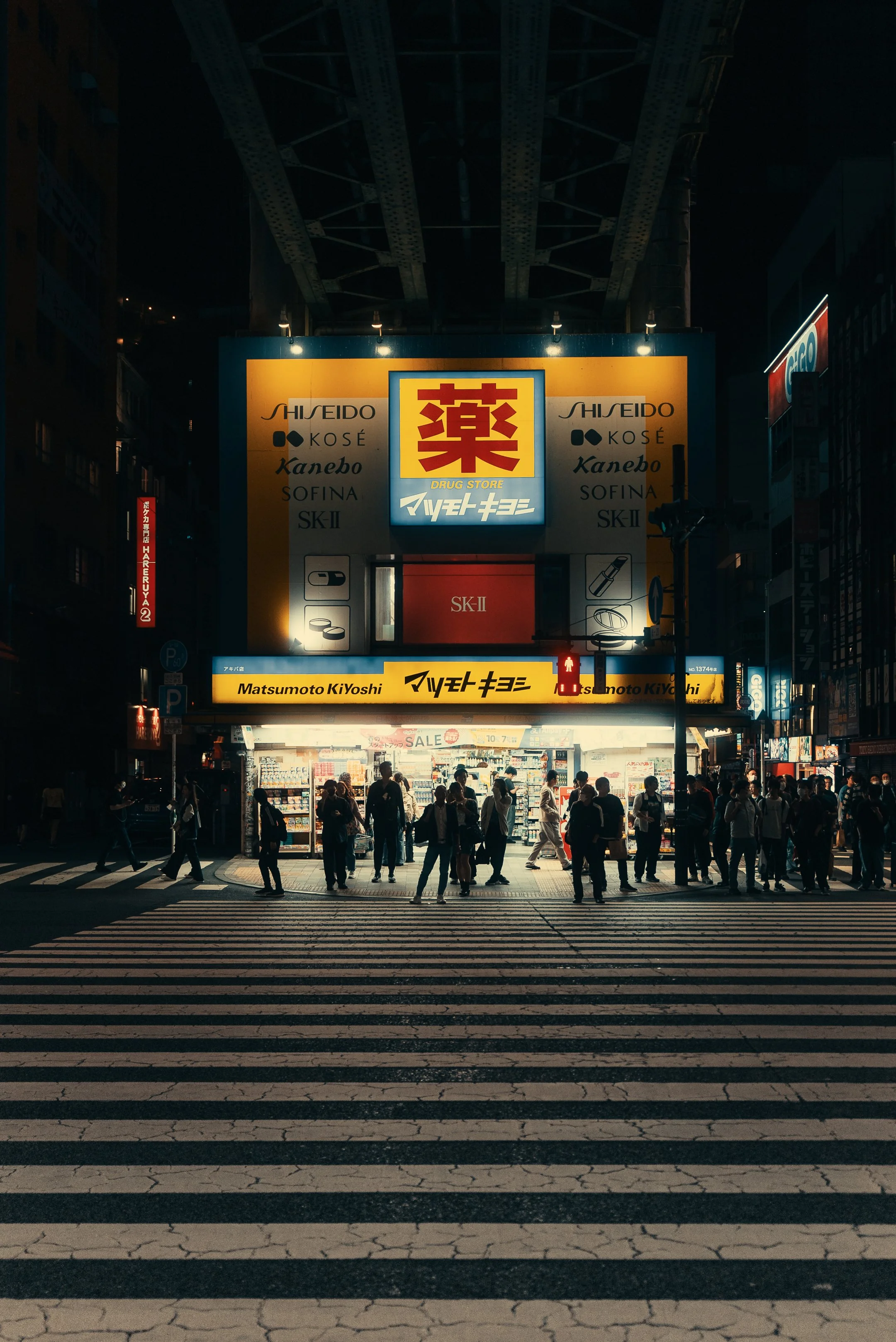 Nighttime busy city street with crosswalk and illuminated store signs, including a large brightly lit sign with red Chinese characters and various smaller signs with Japanese writing. Pedestrians crossing the street, some waiting and some walking.