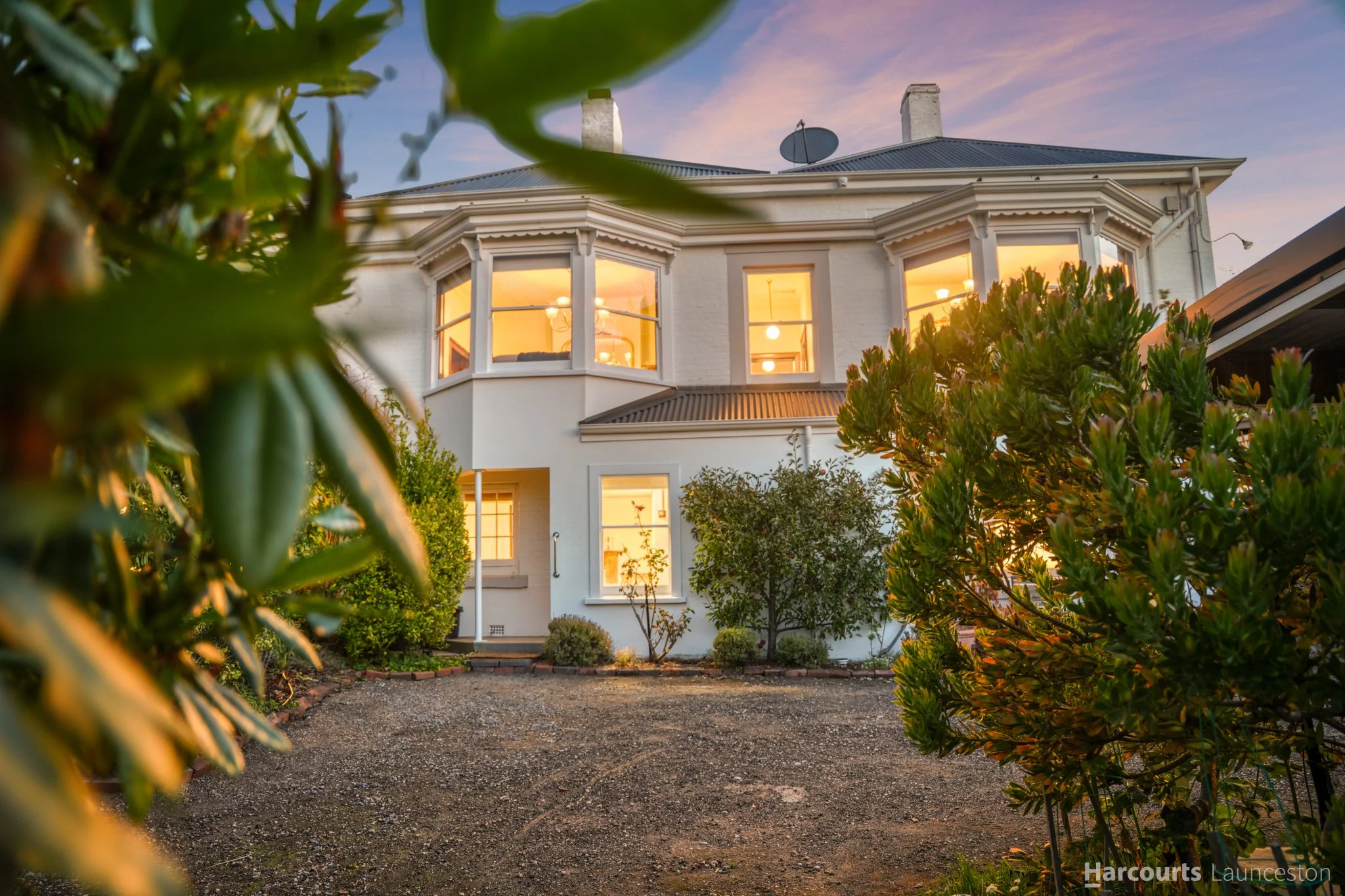 A white house with a dark roof, multiple chimneys, and large lit windows viewed through bushes and trees at sunset.