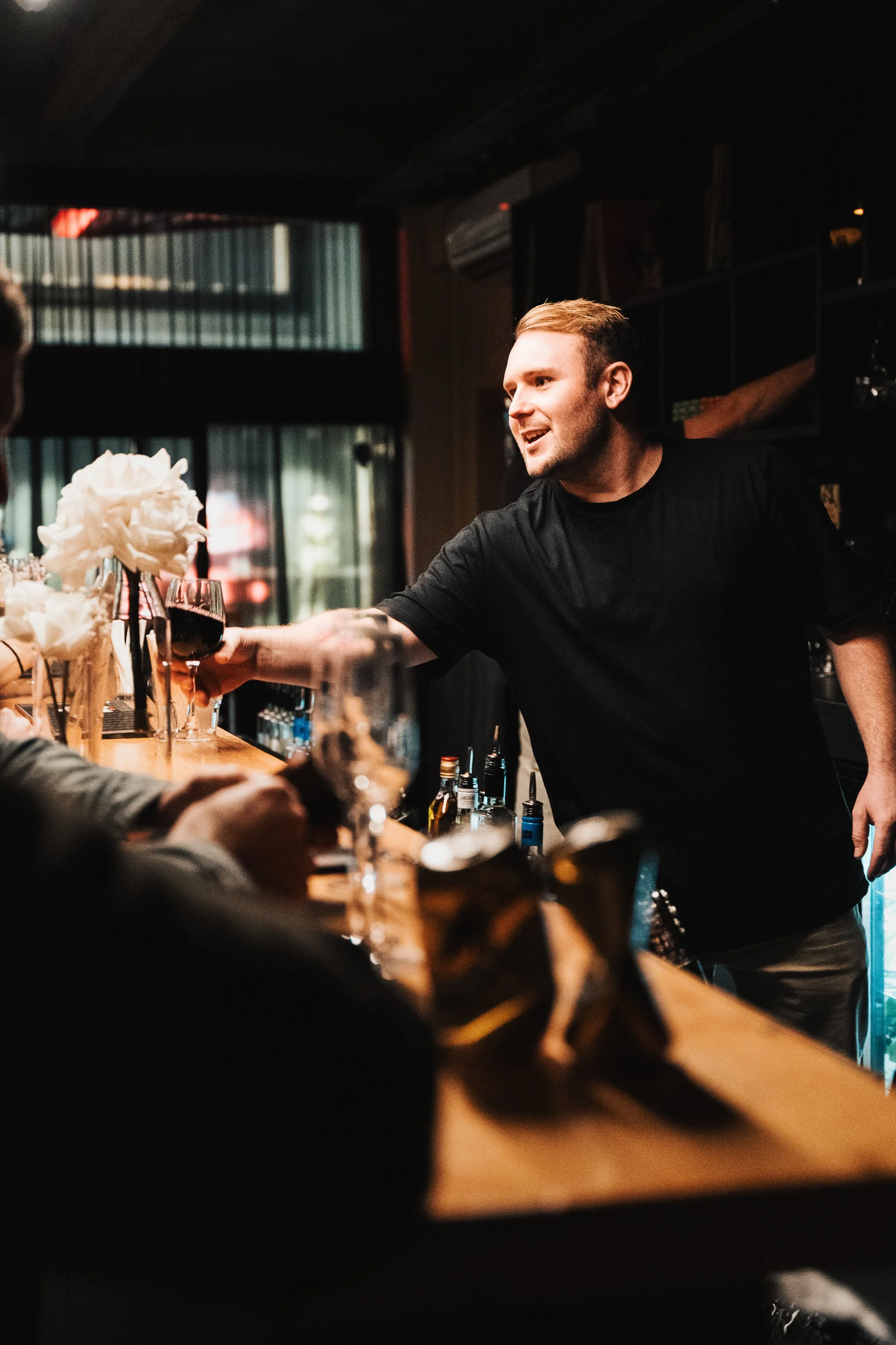 A man with short blond hair and wearing a black T-shirt is smiling and handing a glass of red wine to someone across a bar counter in a dimly lit bar or restaurant.