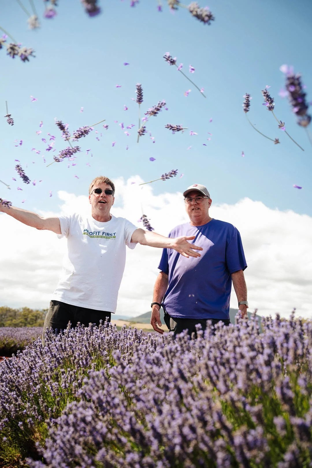 Two men standing in a lavender field with purple lavender flowers in the foreground and background, one man in a white shirt with sunglasses and the other in a blue shirt and baseball cap, with blue sky and clouds above.