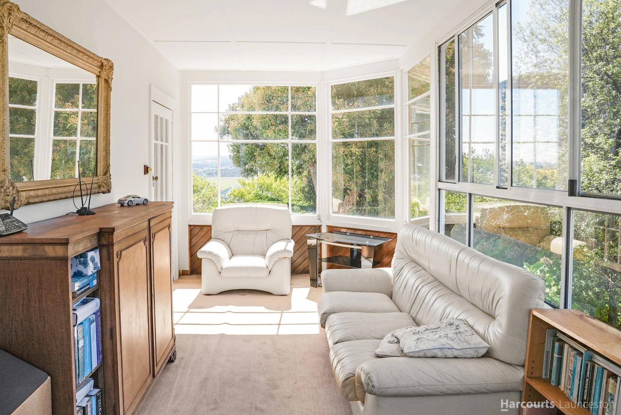 Sunlit living room with large bay windows showing trees outside. There are two white couches, a wooden cabinet with books and a model car on top, and a mirror on the wall.