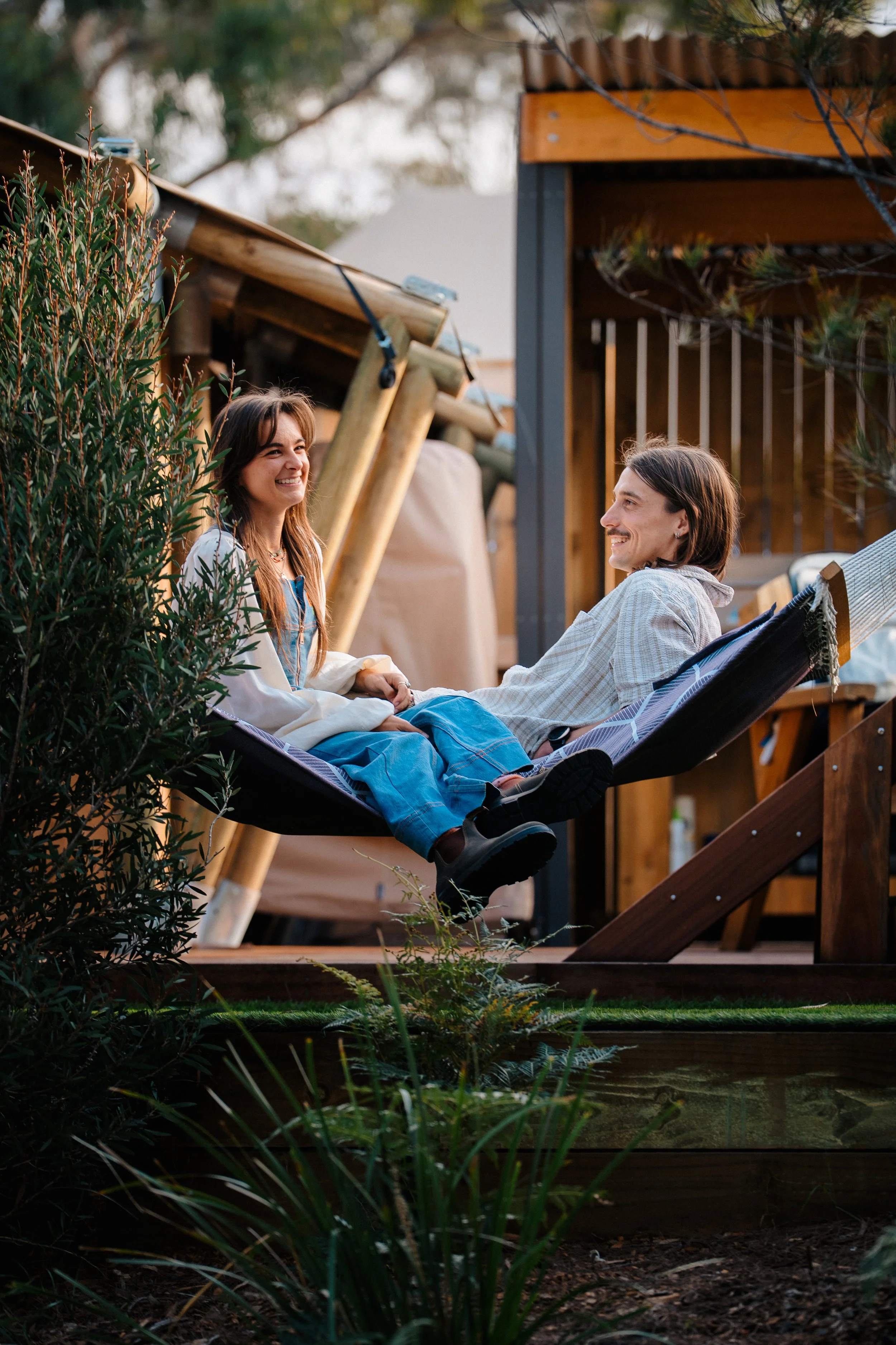 Two people are sitting on a porch swing, smiling at each other. They are surrounded by outdoor plants and a wooden structure in the background, with trees visible in the distance. The setting appears to be a backyard or garden.