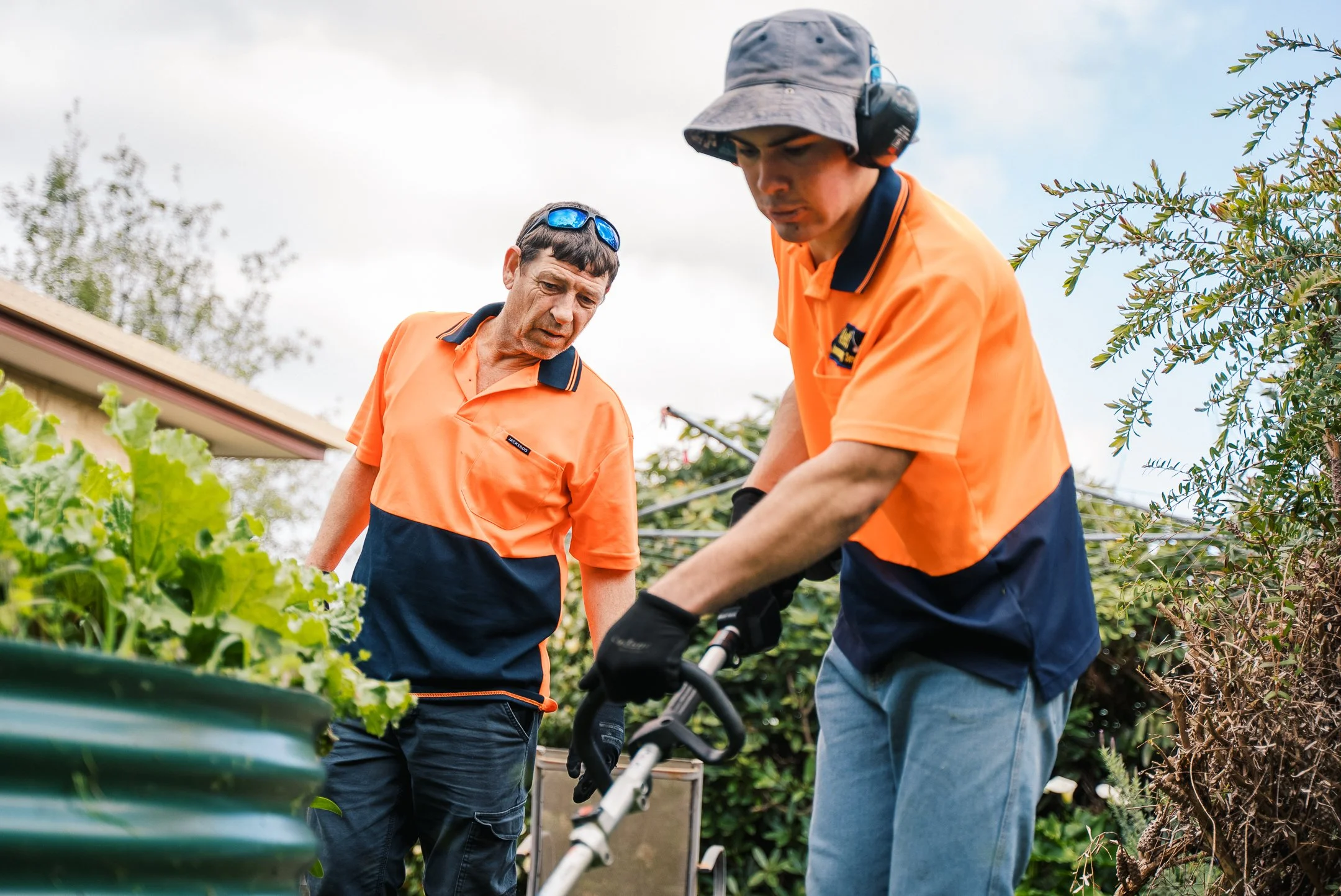 Two men wearing orange and navy work uniforms using gardening tools in a garden with plants and trees under a cloudy sky.