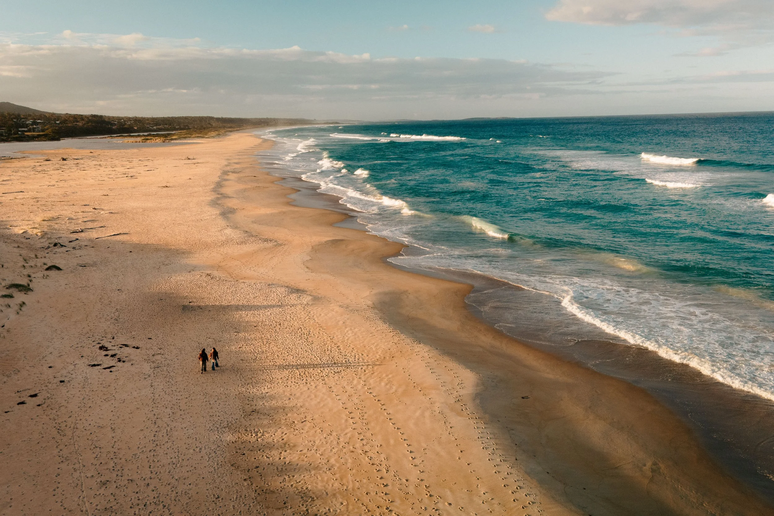 A sandy beach with a few people walking, gentle waves in the ocean, and a distant view of houses and hills under a partly cloudy sky.