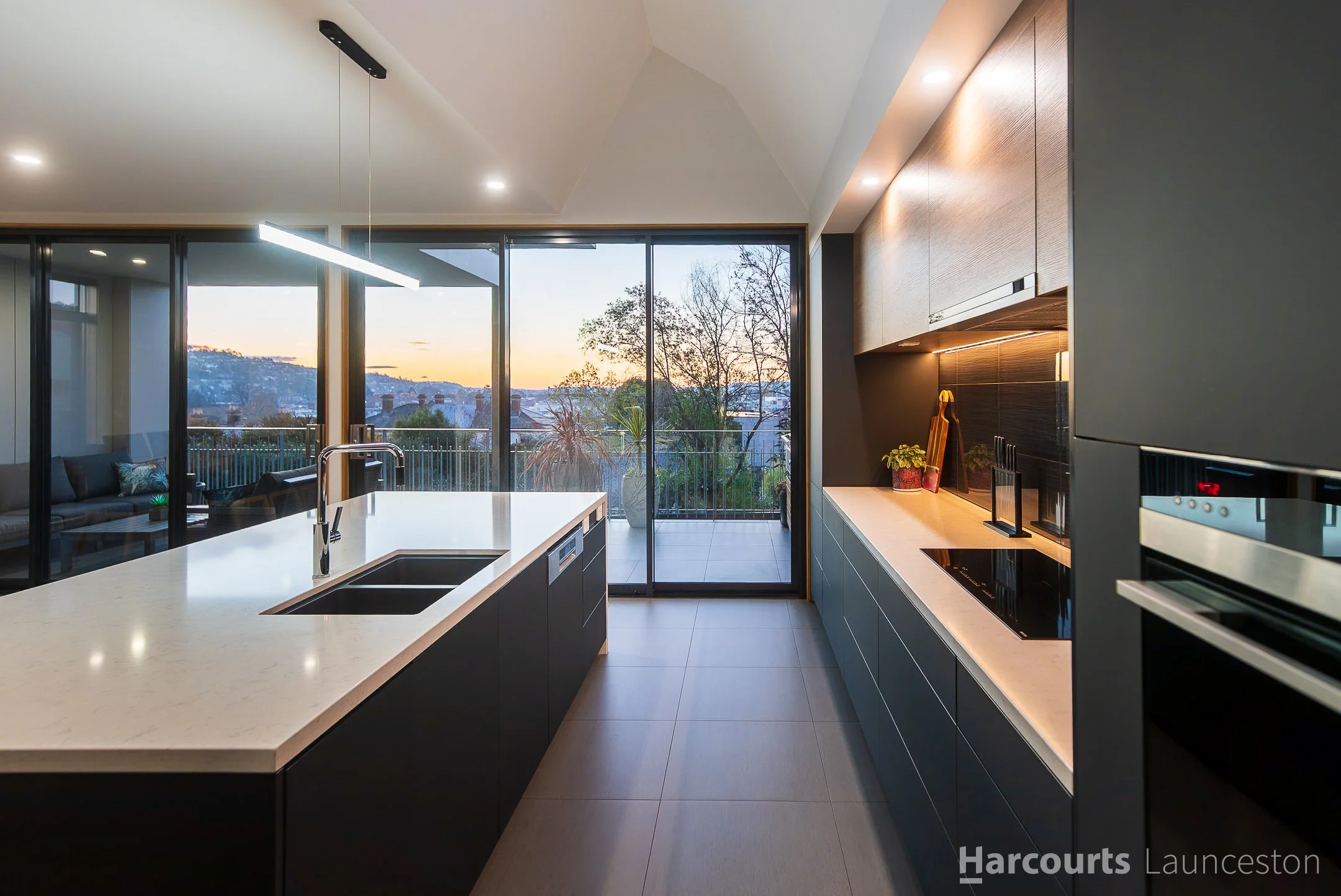 Modern kitchen with a white island, black cabinetry, a large window revealing a scenic view of hills and trees at sunset, and a balcony outside.