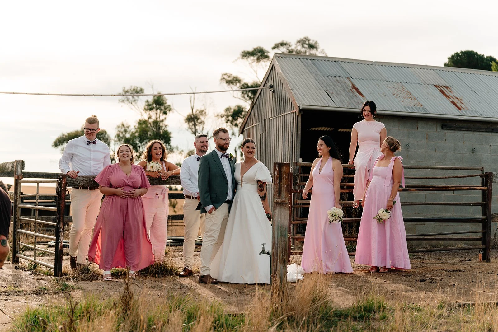 A group of people, including a bride and groom, in wedding attire standing outside a rustic farm building, with bridesmaids and groomsmen smiling and interacting.
