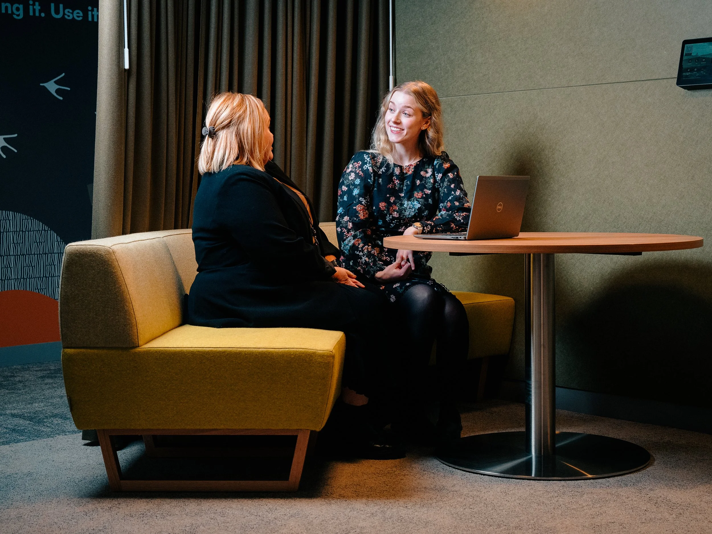 Two women sitting and talking in a conference room near a table with a laptop. One woman has hair tied back with a clip, and the other has curly blonde hair, wearing a floral dress. There are curtains and a dark wall in the background.