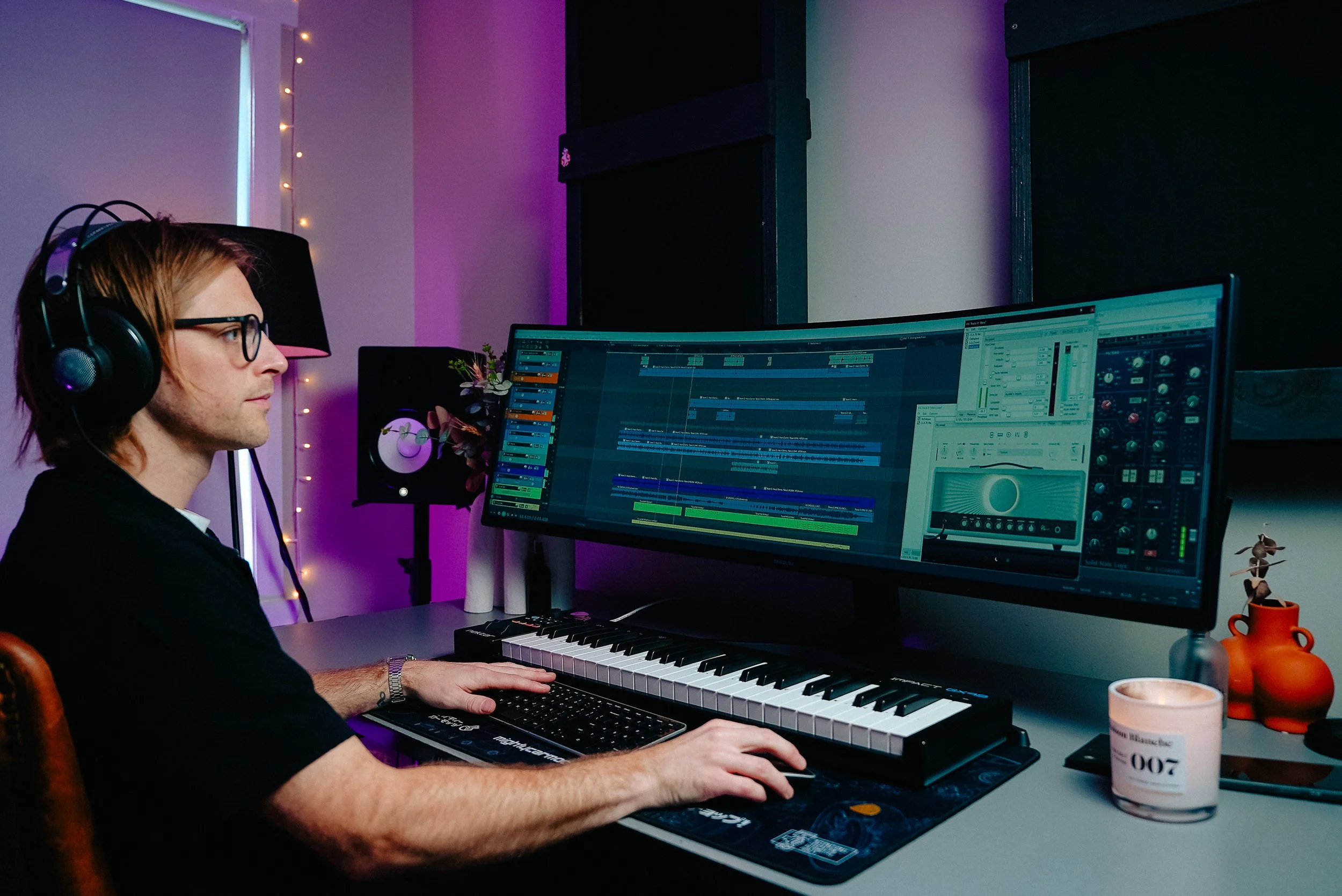 A person with red hair and glasses wearing headphones working on music production on a large dual-monitor setup with a keyboard, mouse, and a MIDI controller on the desk. The room has colorful lighting, fairy lights, and various decorative items incl