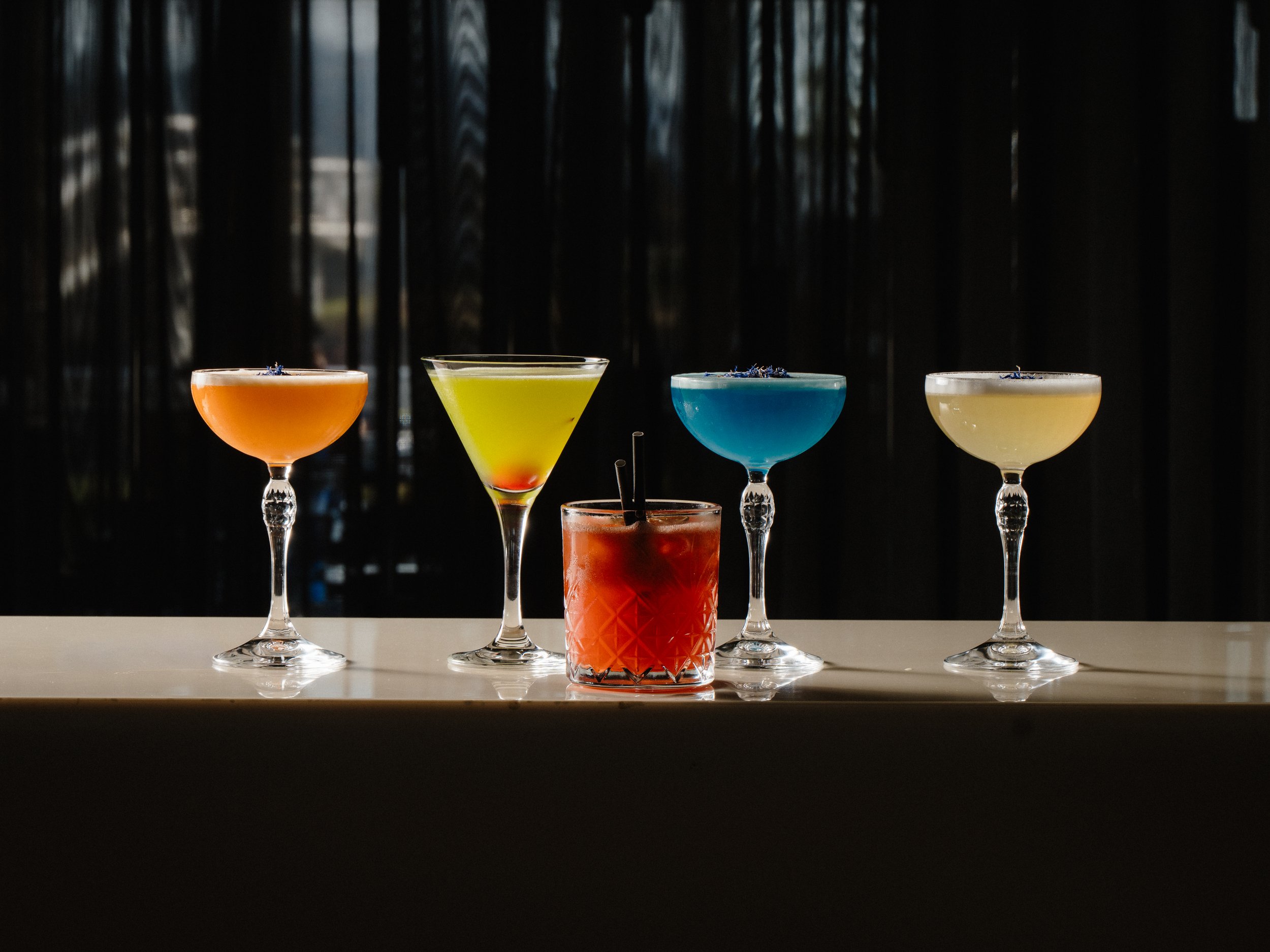 Five colorful cocktails in elegant glassware lined up on a white surface with a dark, blurred background.