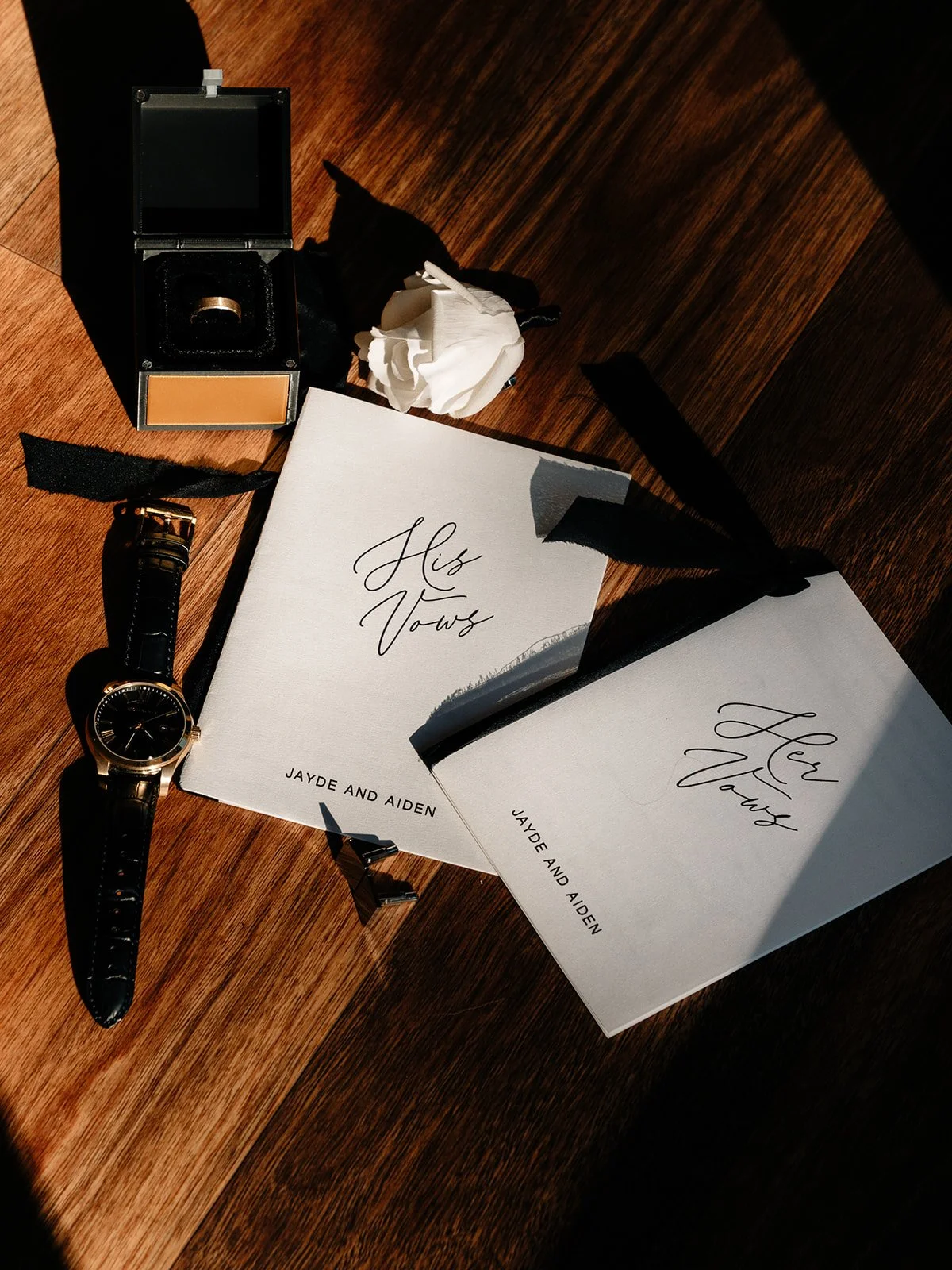 Wedding invitation cards that say "His Vows" and "Her Vows" with the names Jayde and Aiden, a wristwatch, a black ring box, and a white flower on a wooden surface.
