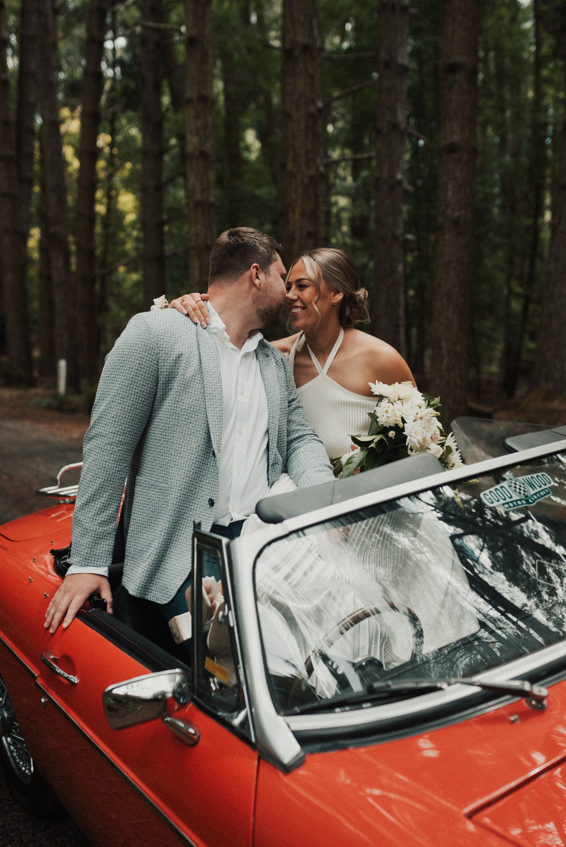 A bride and groom sharing a kiss inside a red convertible car in a forested area during daytime.