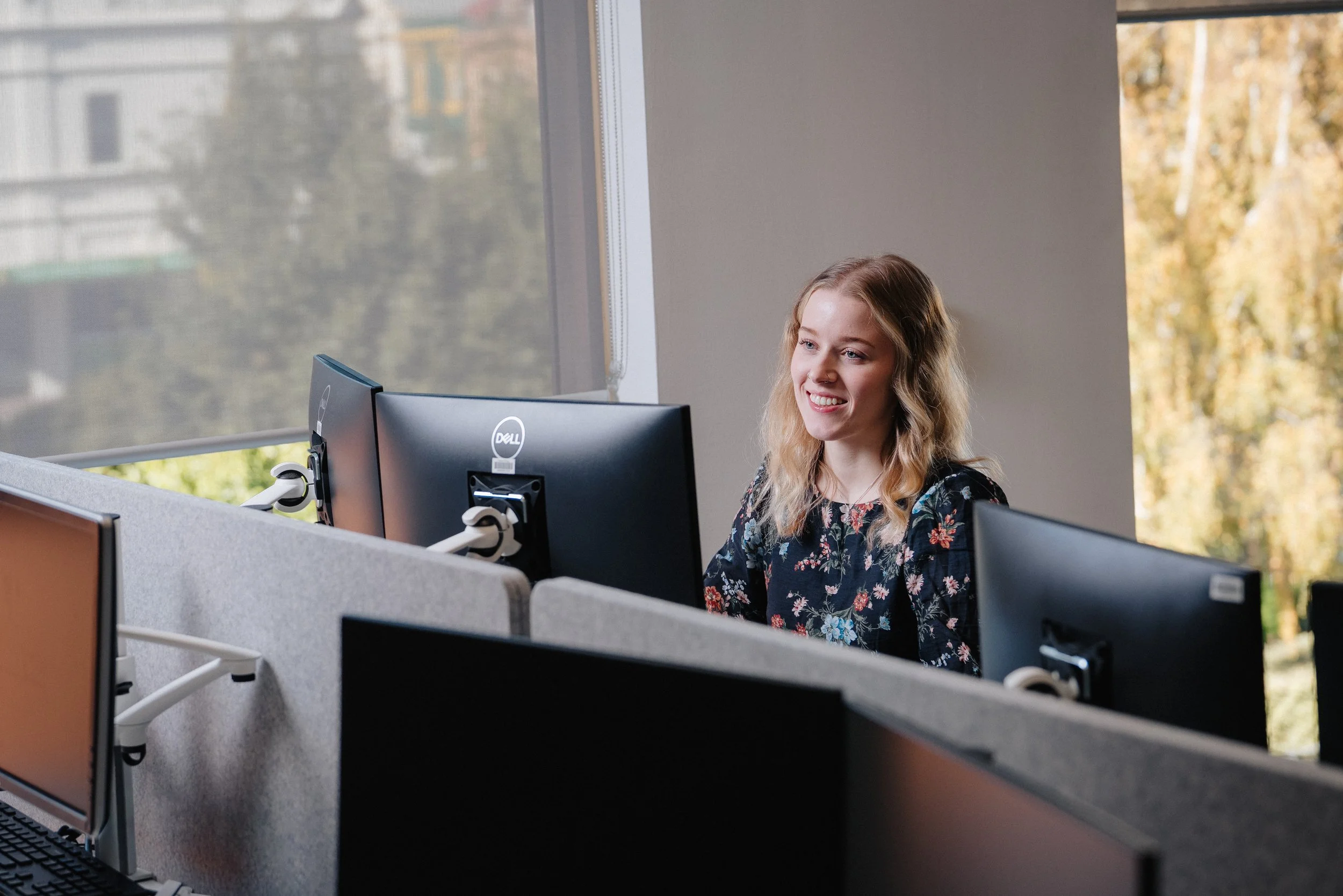 A woman with long blonde hair and a floral top working at a computer in an office with large windows showing trees and buildings outside.