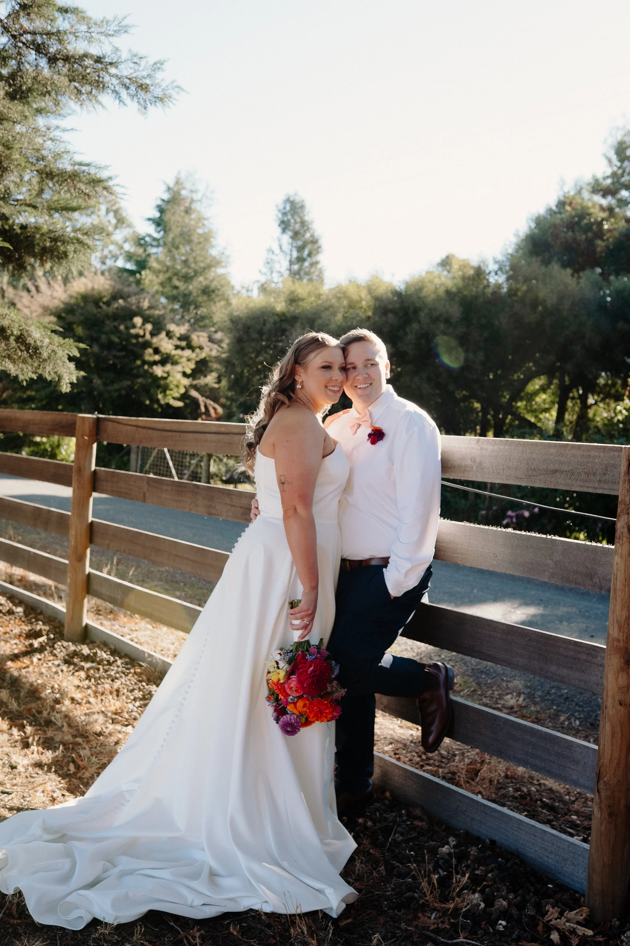 A bride and groom pose together outdoors near a wooden fence, with trees and clear sky in the background. The bride is holding a bouquet of vibrant flowers, and they are smiling closely together, dressed in wedding attire.