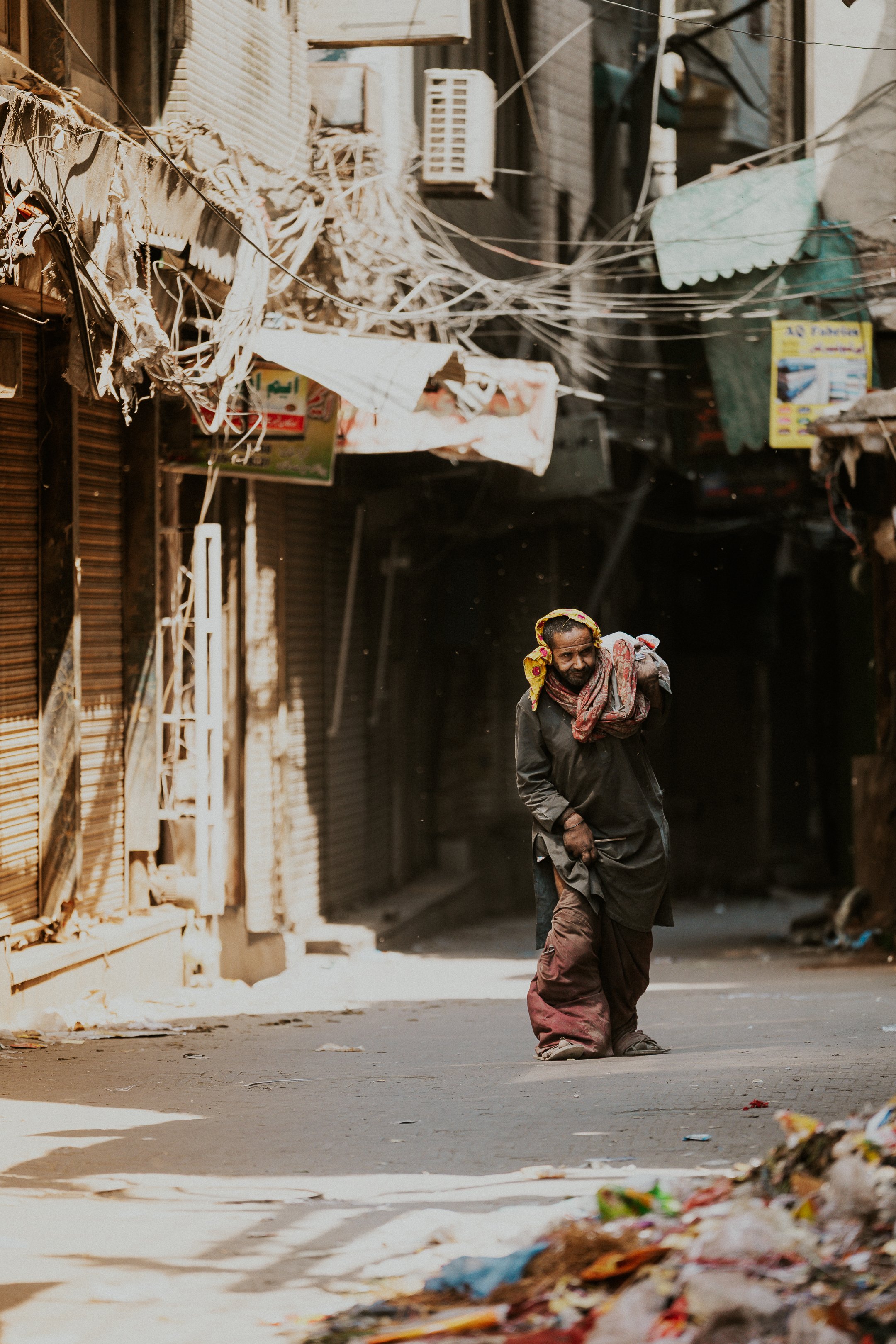 A man walking on a street with closed shops and electrical wires overhead, surrounded by trash and debris.