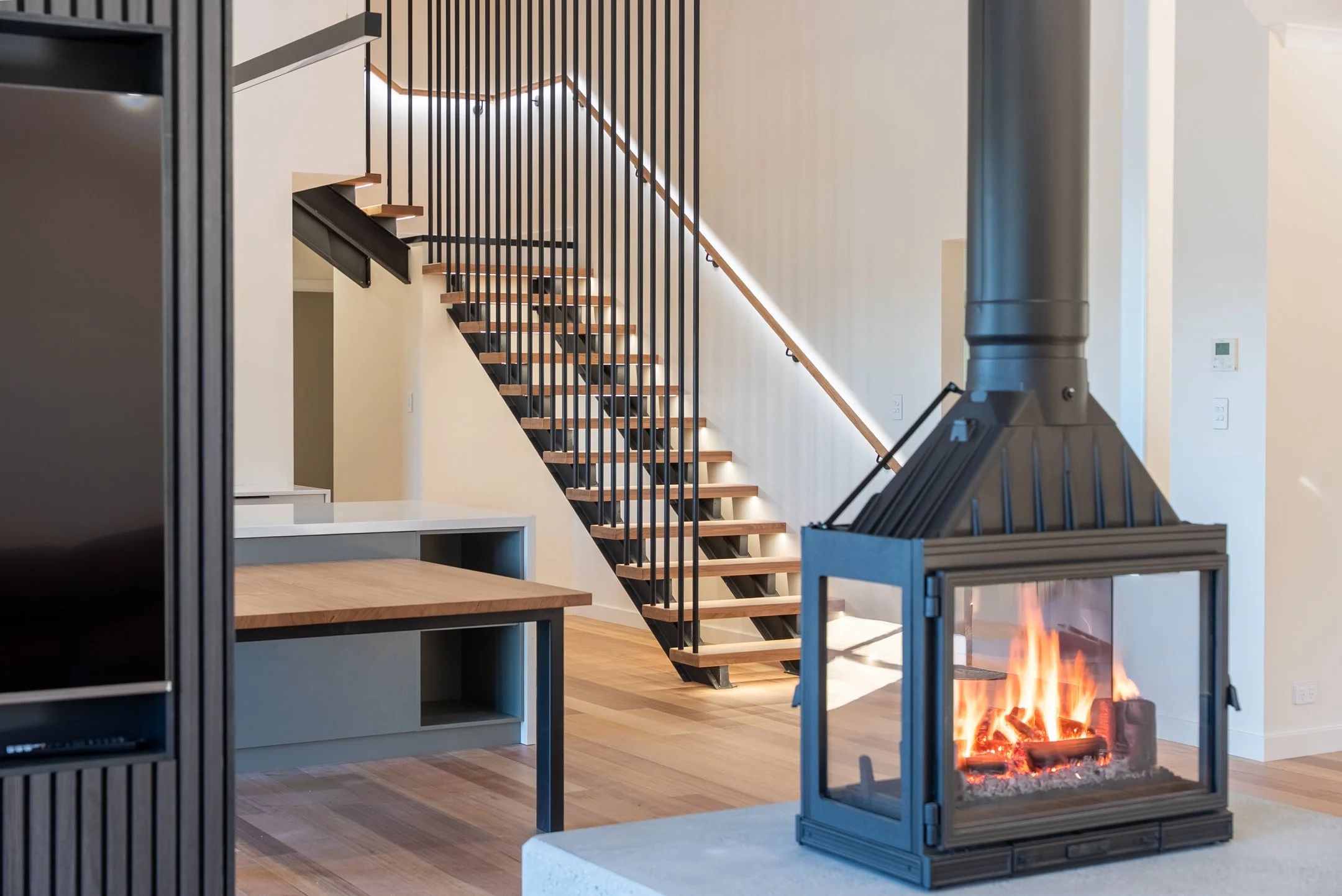 Interior view of a modern living room with a staircase, a fireplace, and shelves. The staircase has wooden steps and black metal railing. The fireplace has a glass front with visible flames. The room has light-colored walls and wooden flooring.