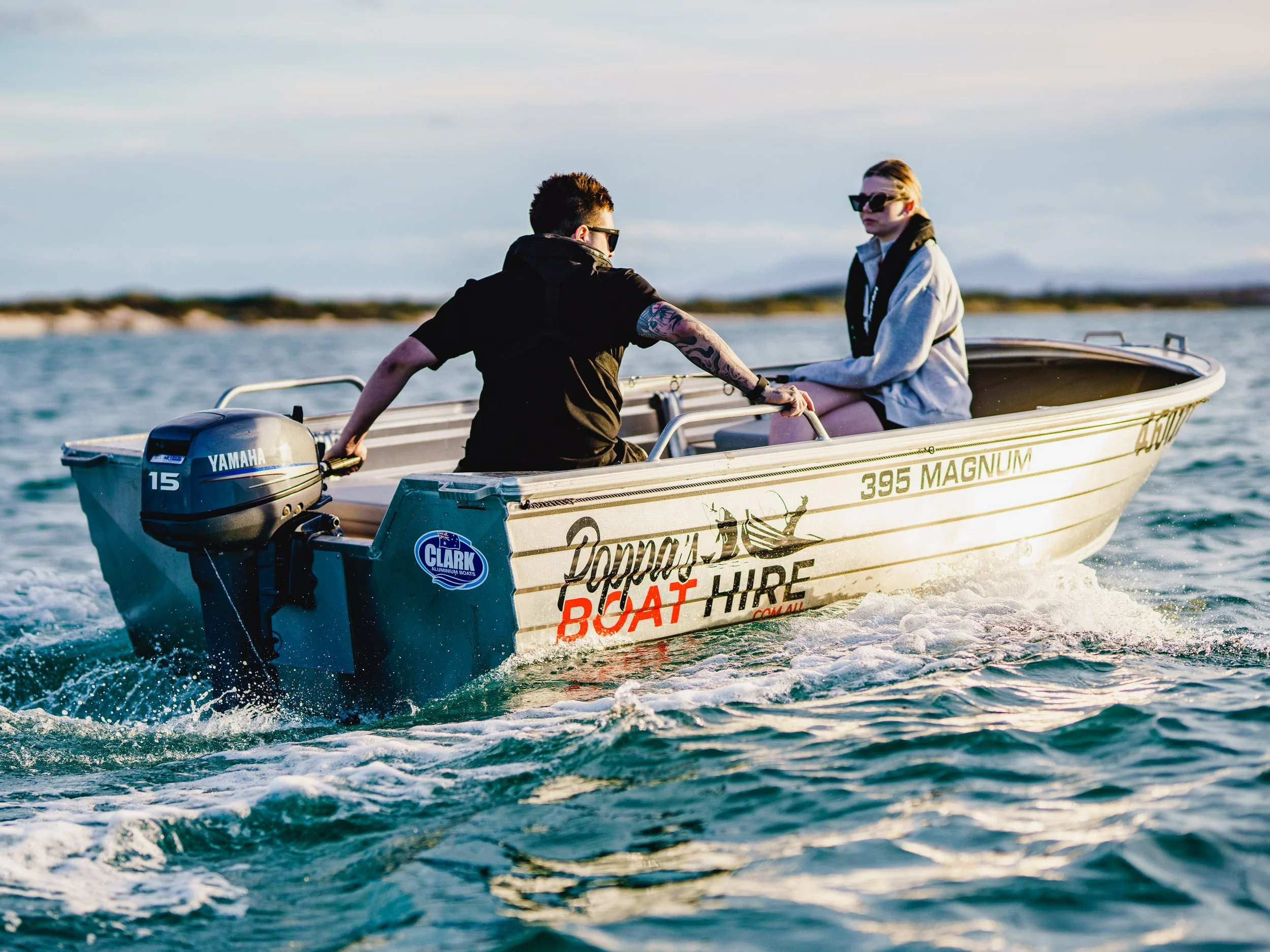 Two people are sitting in a small boat on the water, with a Yamaha outboard motor. They are wearing sunglasses and clothing suitable for a mild weather day. The boat has branding including 'Poppy's Boat Hire' and is moving through the water with a bl