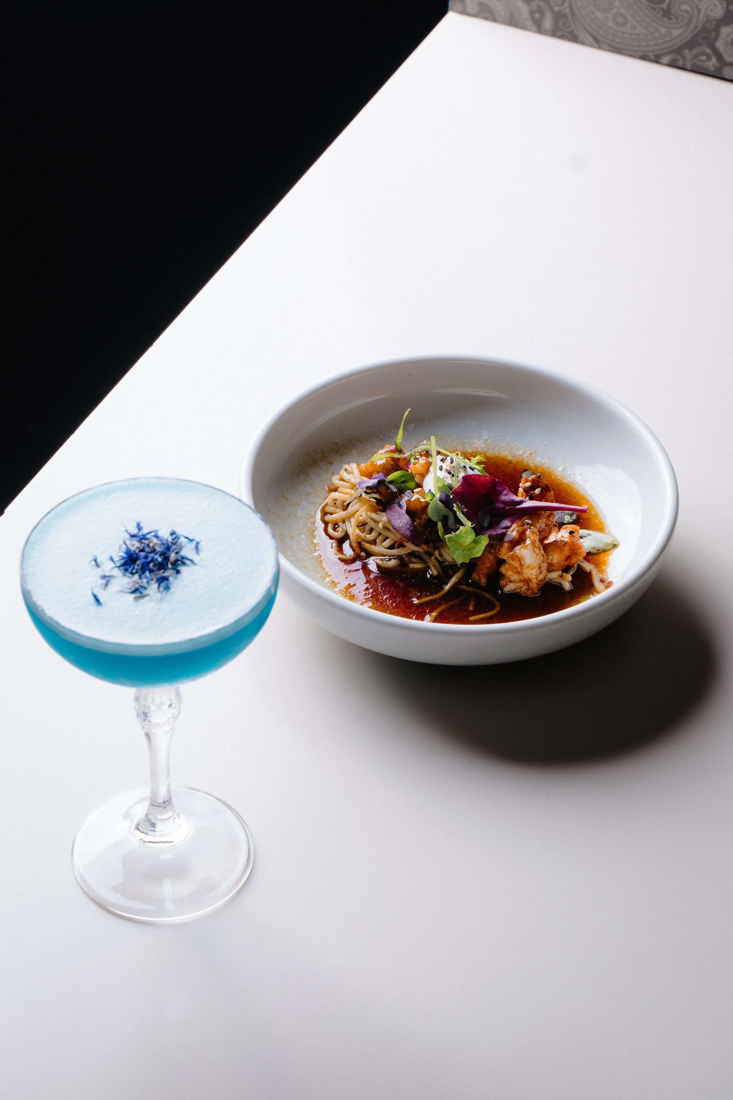 A glass of blue cocktail with blue edible flowers and a white bowl of Asian-style noodle soup topped with greens and herbs, on a white surface against a black background.
