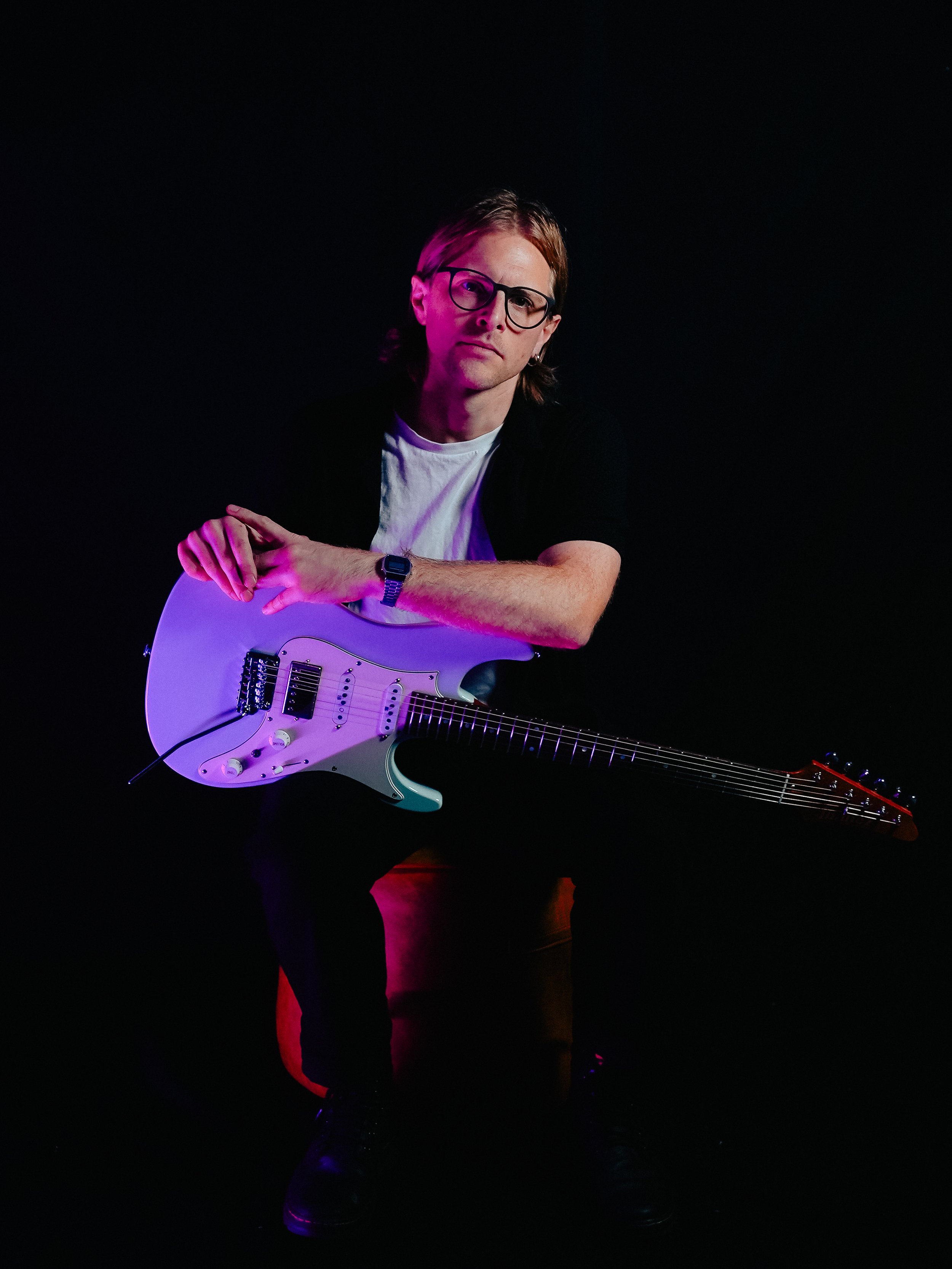Man with glasses holding a white electric guitar against a black background with pink and red lighting highlights.