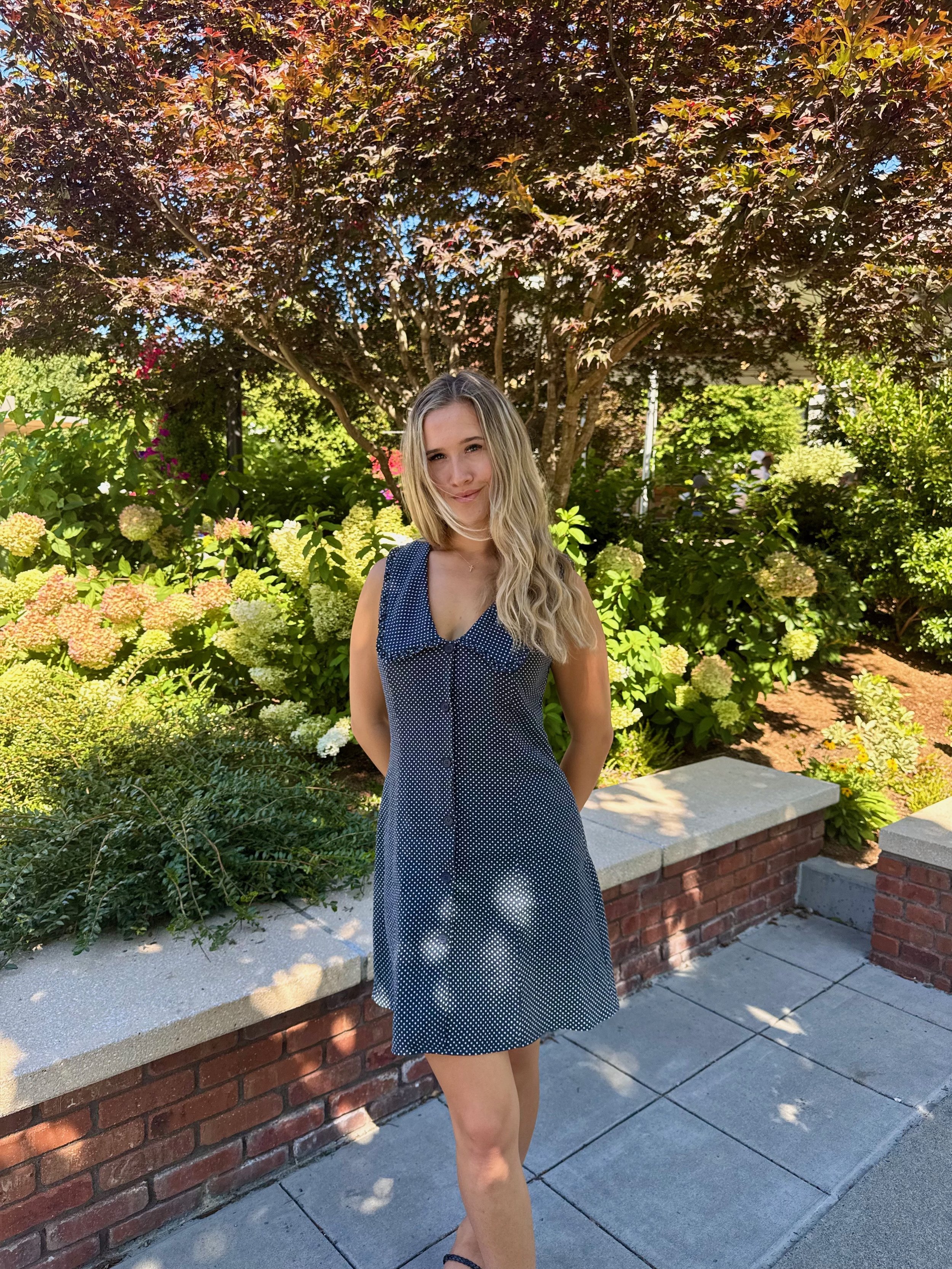 A young woman with long blonde hair and light skin is standing outdoors in front of a large leafy tree and various flowering plants. She is wearing a sleeveless navy blue dress with white polka dots and has a subtle smile. The scene is sunny, and she is standing on a paved sidewalk next to a low brick wall with a concrete top.