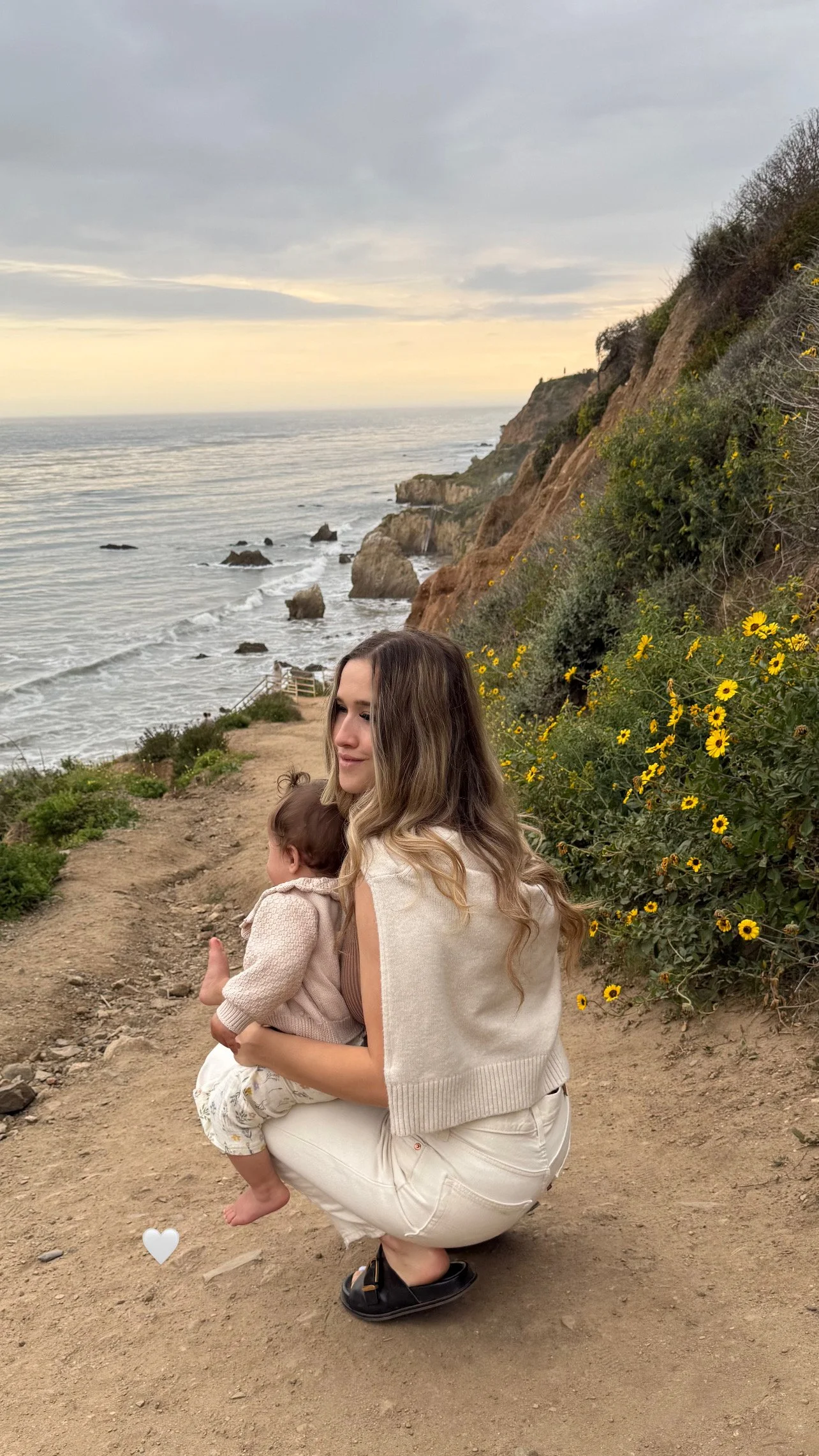 A young woman crouching on a dirt path holding a small child with long brown hair near a scenic coastline with rocky cliffs, yellow flowers, and the ocean in the background during sunset.