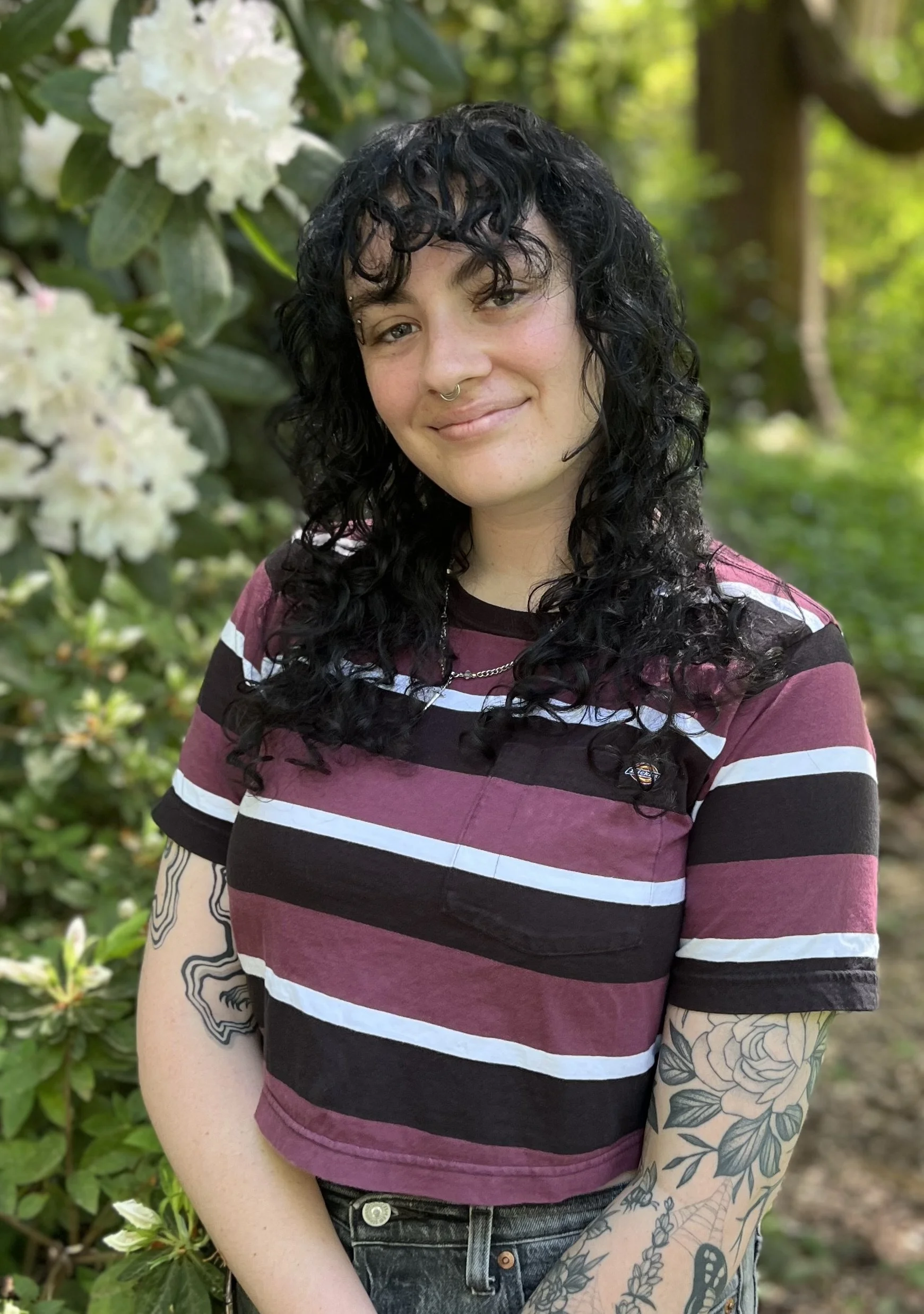 A person with black curly hair, tattoos on the arms, and a septum piercing smiling outdoors near white flowers and green foliage.
