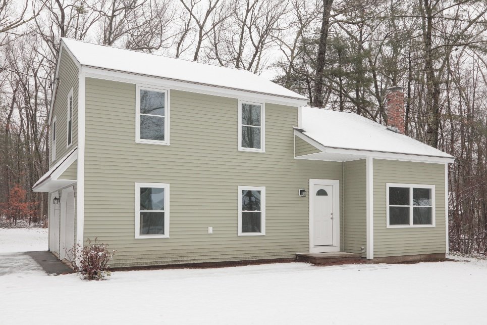 A two-story house with beige siding, white trim, and a white front door, set against a snowy landscape with leafless trees in the background.