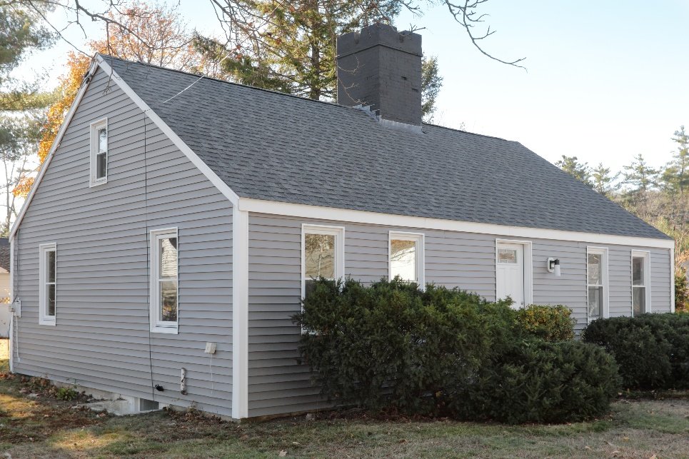 A gray house with white trim, a dark chimney, and multiple windows, surrounded by bushes and trees with clear sky in the background.