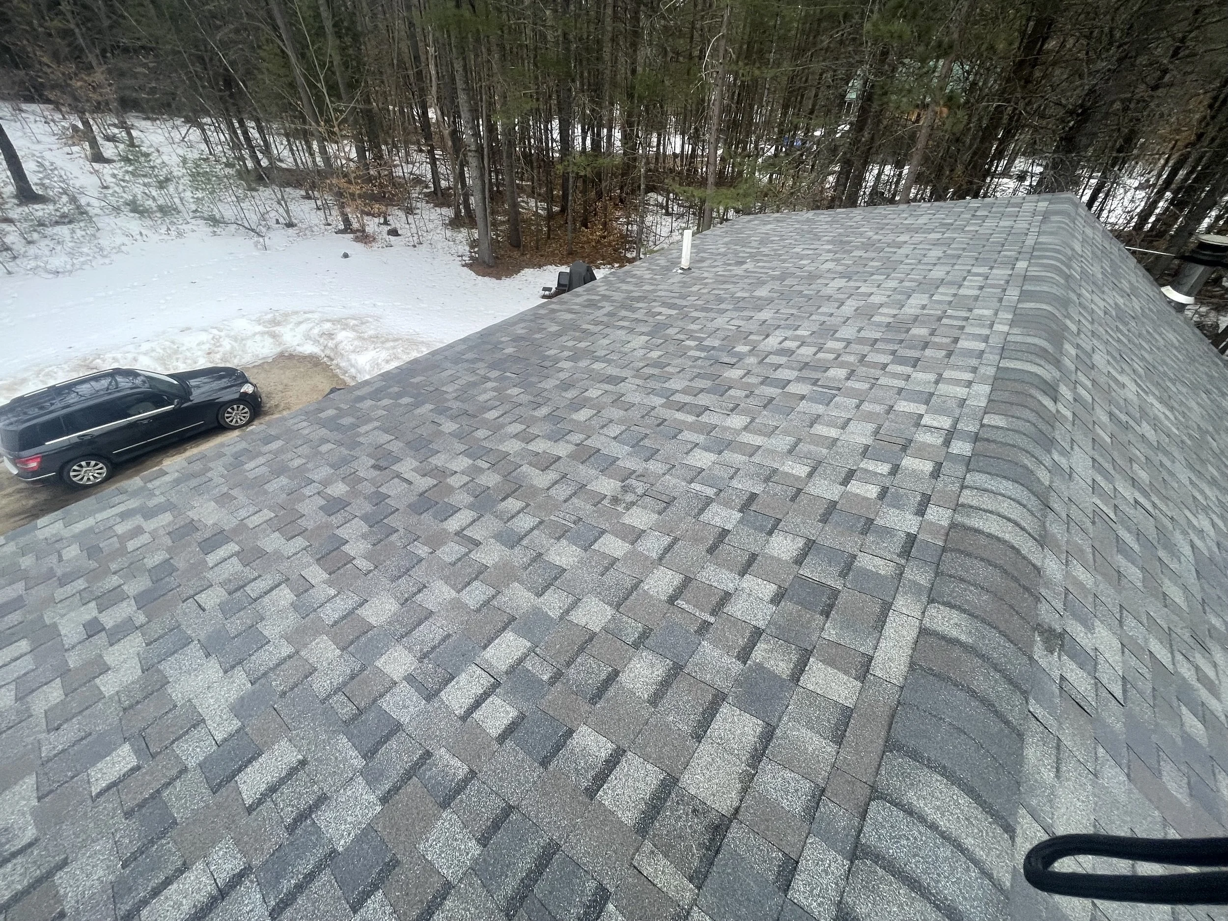 View of the roof of a house with shingles, residential area with trees and snow-covered ground visible in the background.