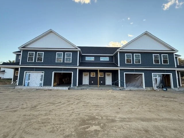 New two-story residential building under construction with unfinished garage spaces, set on a dirt lot, under a partly cloudy sky.