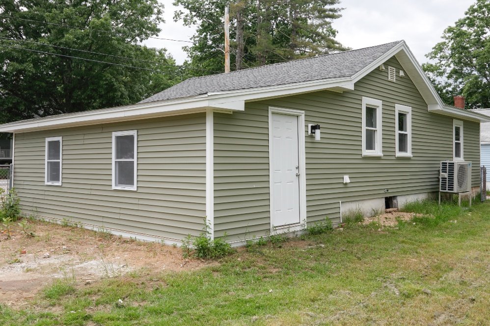 A single-story house with light green vinyl siding, white trim, and a grey shingled roof. It has four windows, a white door, and an air conditioning unit outside. The house is surrounded by grass and some dirt patches.