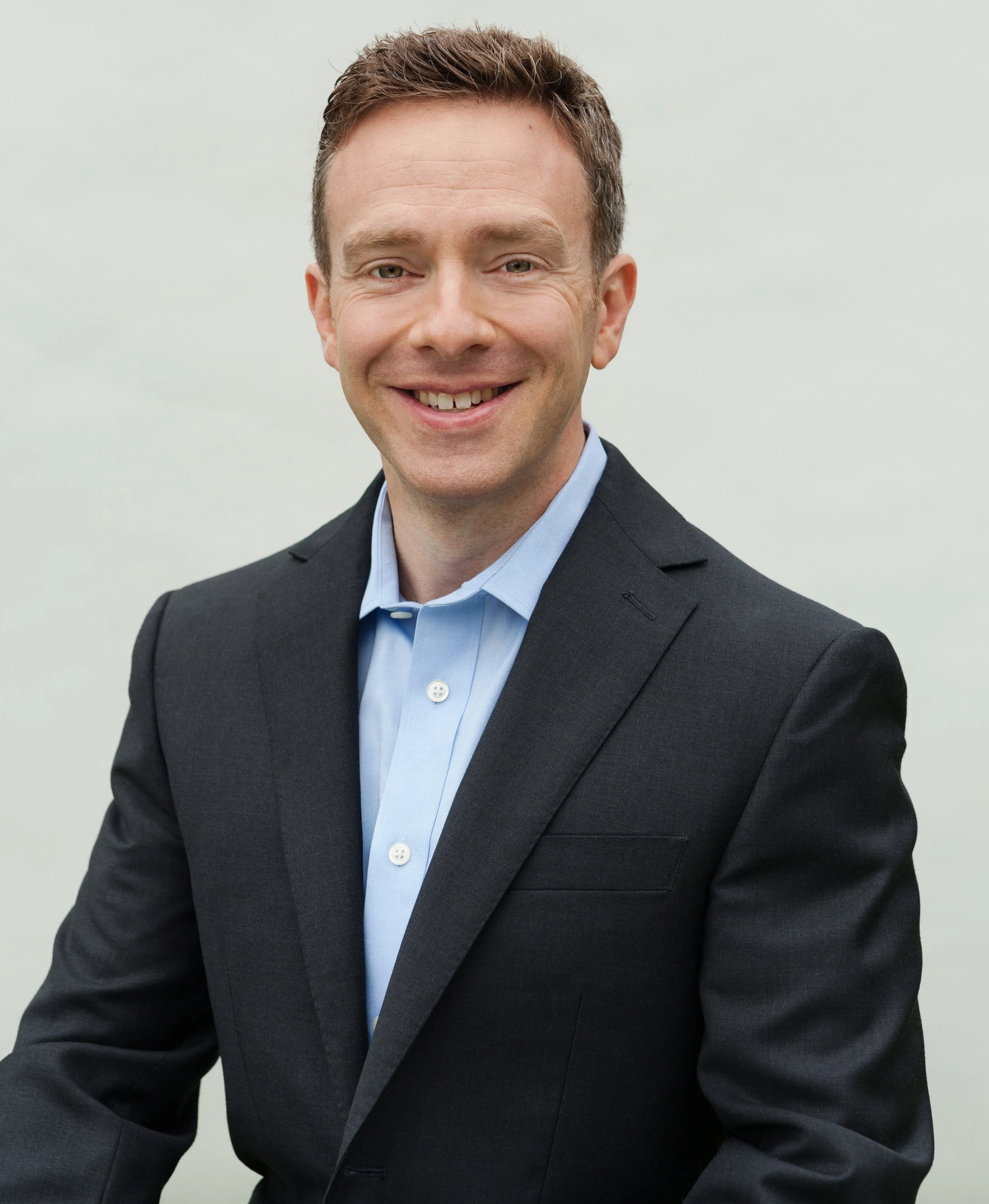 A smiling man with short brown hair, wearing a dark suit jacket and a light blue dress shirt, against a plain light background.