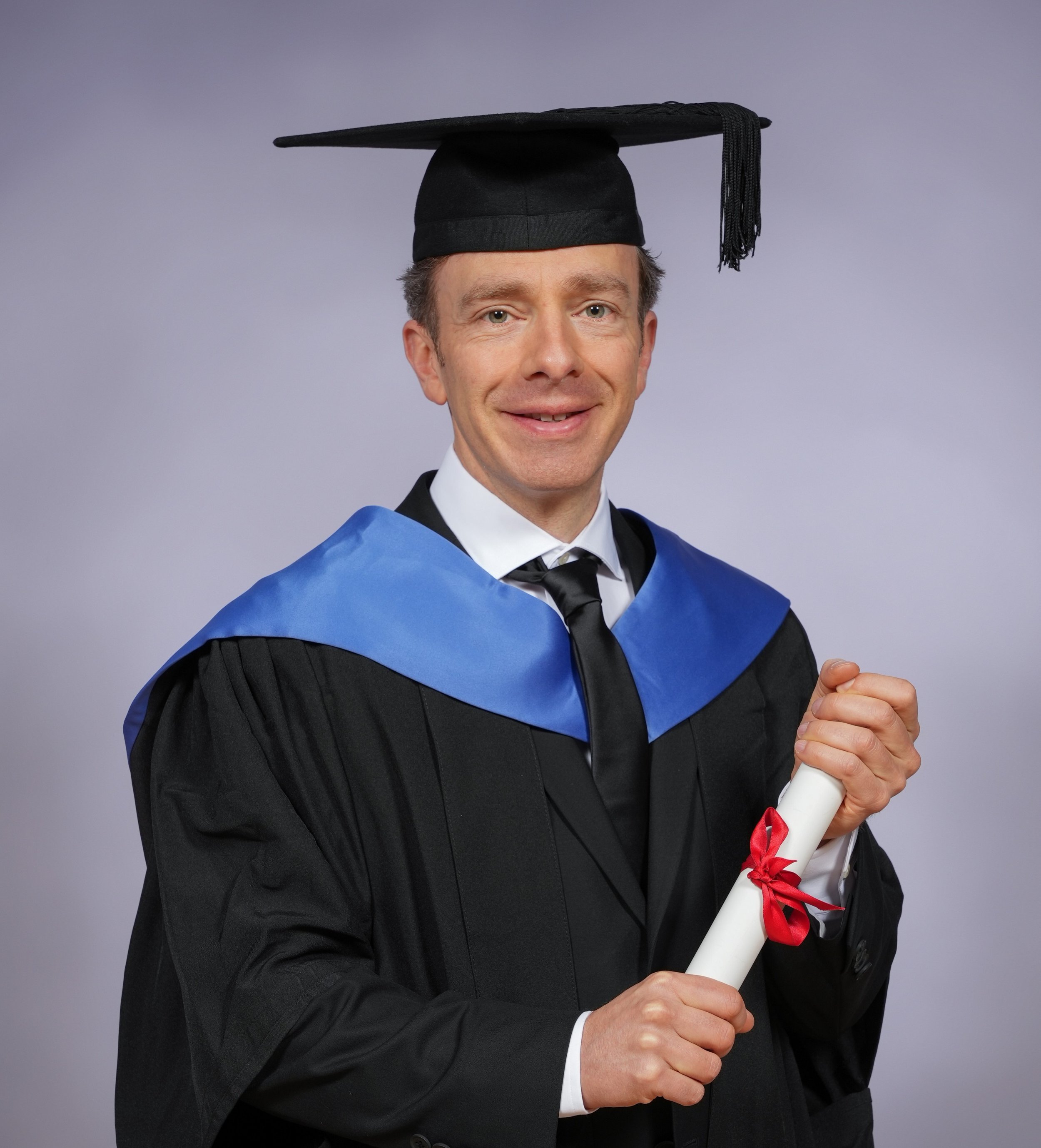 A man in a graduation cap and gown holding a diploma tied with a red ribbon.