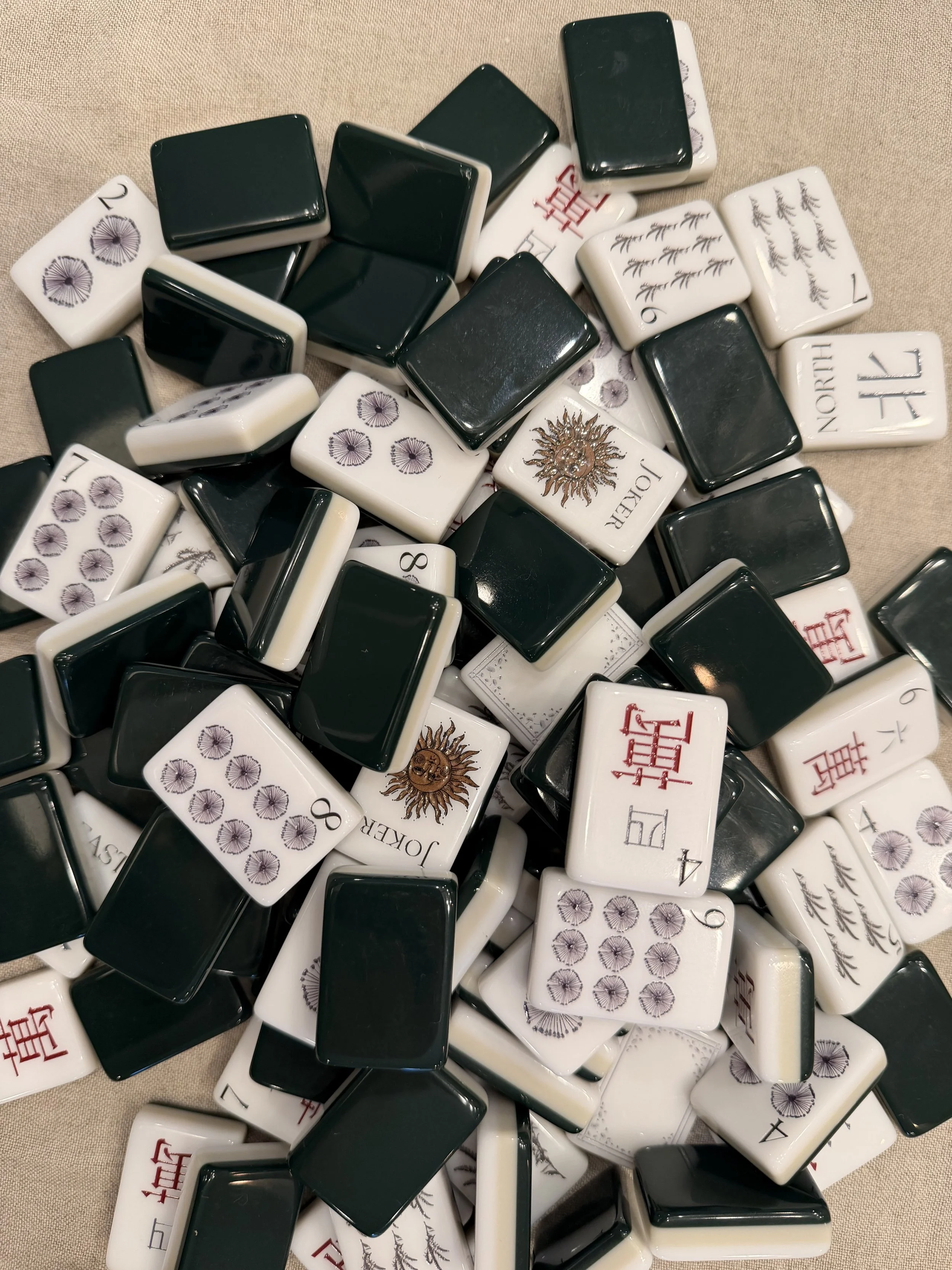 Overhead view of Winter Solstice tiles heaped on linen, some showing deep green backs and others showing faces with native dandelion Dots, red Cracks, grass Bams, and gold sun Jokers.