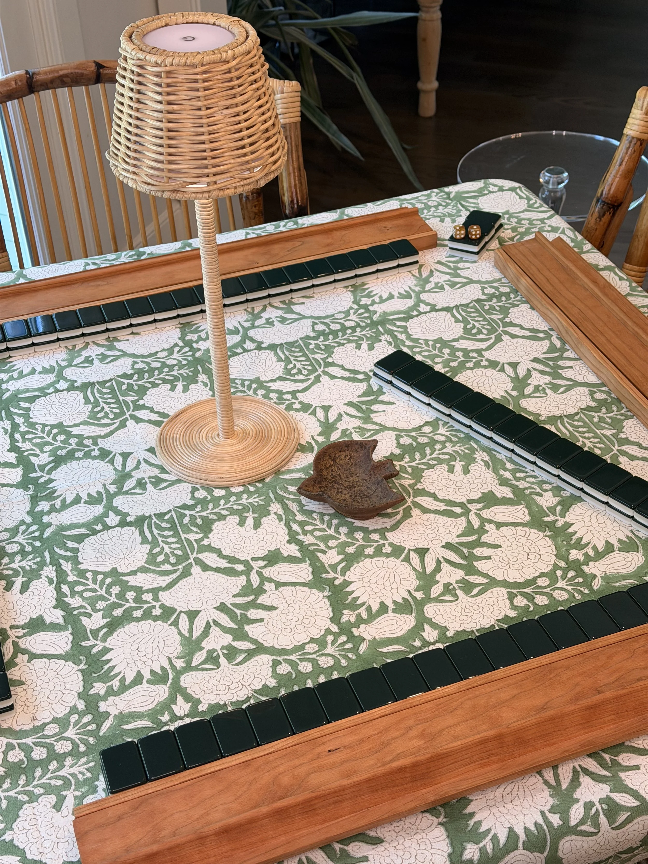 A mahjong table set for play: walls of green-backed Winter Solstice tiles inside four wooden racks on a green-and-white floral tablecloth, with a wicker lamp and gold dice.